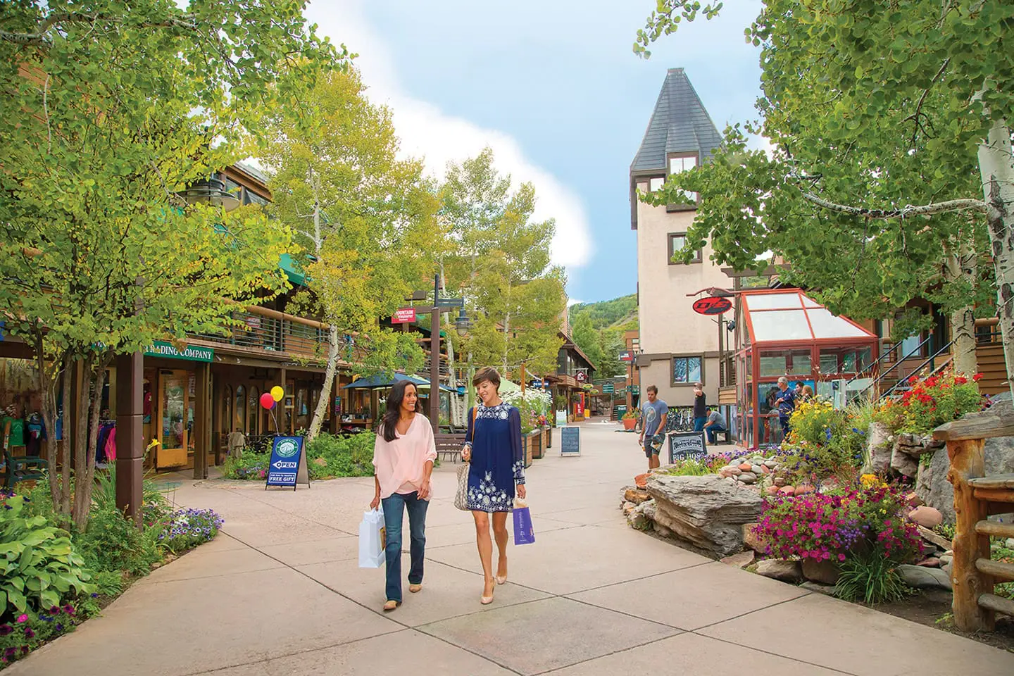 This image showcases a charming, pedestrian-friendly shopping plaza featuring rustic architecture, lush greenery, and blooming flower beds. Two women are walking along the paved pathway, which is flanked by shops, restaurants, and a distinct mountain-style watchtower, conveying a welcoming and vibrant resort community atmosphere.