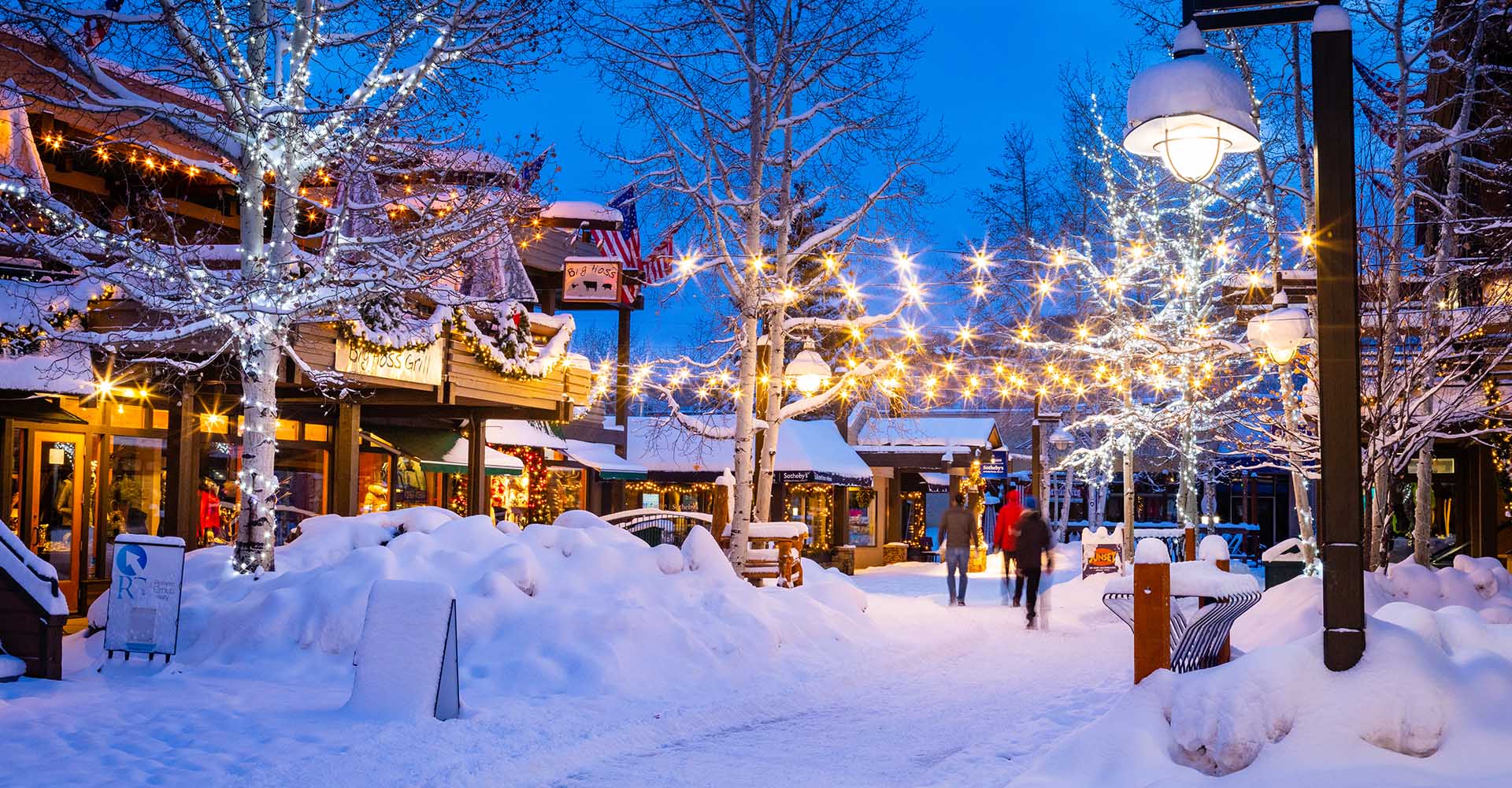 This picturesque image captures a charming, snow-covered pedestrian walkway in an upscale mountain village setting at twilight. The scene is illuminated by glowing string lights draped through trees and classic street lamps, highlighting boutique storefronts and eateries like Big Hoss Grill. The festive, inviting atmosphere suggests a high-end resort town, perfect for seasonal tourism and community engagement.