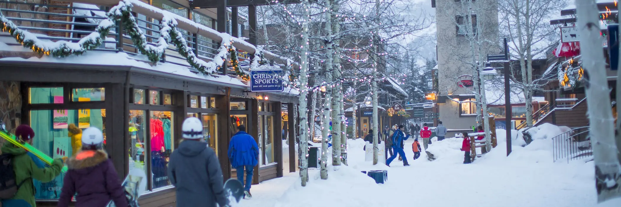 This image captures a charming, snow-covered pedestrian walkway in a picturesque ski village, lined with retail storefronts decorated for the holiday season. The area features a rustic architectural style, with wooden structures, festive lighting on trees, and a vibrant atmosphere with visitors navigating the snow-dusted village center. It showcases a desirable, high-traffic commercial setting perfect for mountain resort tourism.