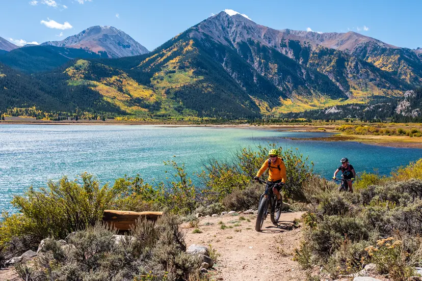 This image showcases a scenic outdoor recreation area featuring a dirt mountain biking trail that runs alongside a pristine lake. In the background, majestic mountains with seasonal autumn foliage frame the tranquil landscape, highlighting excellent local recreational amenities. The setting suggests an active, nature-focused community lifestyle with access to beautiful natural surroundings.