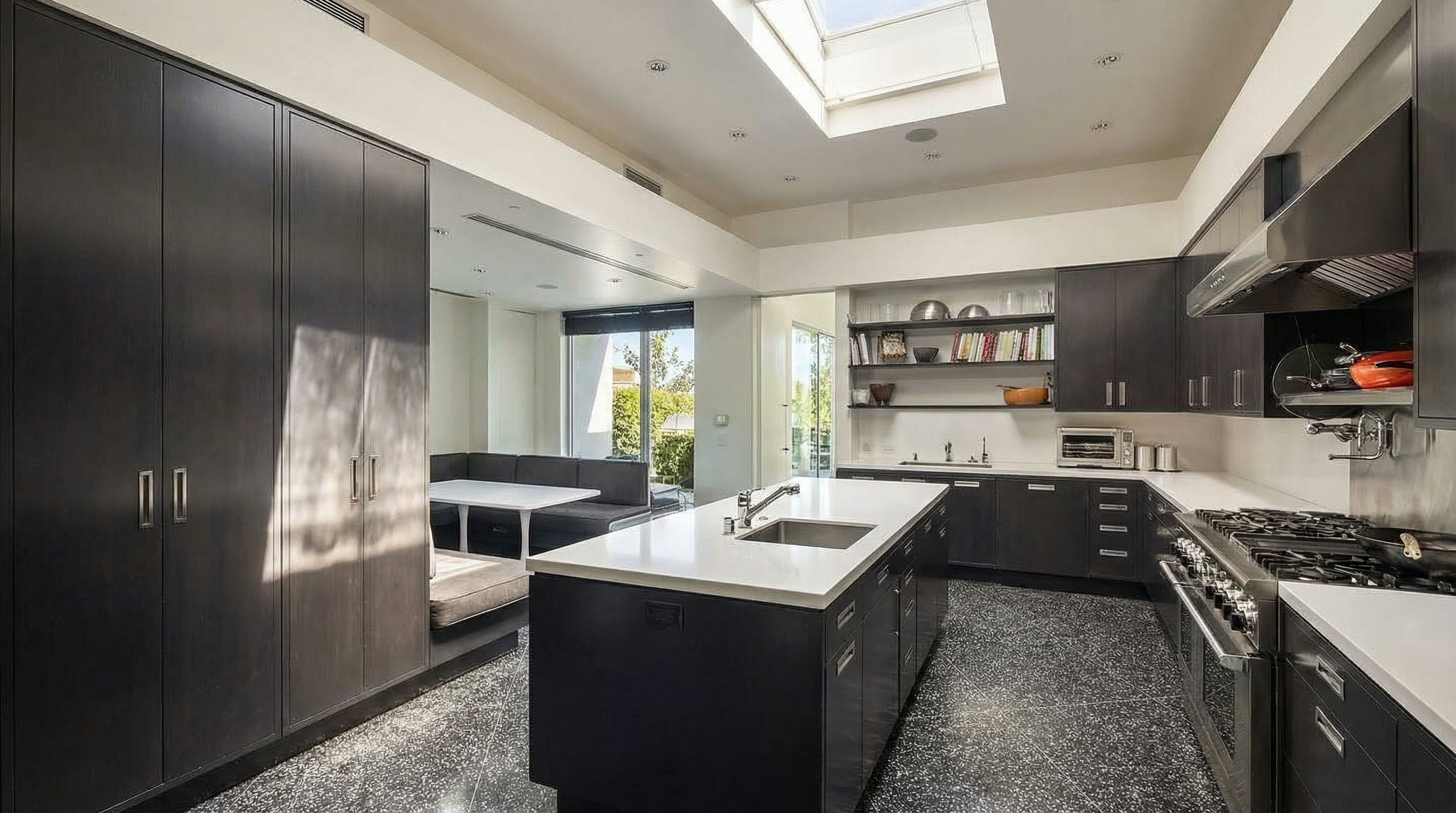 A modern kitchen features dark wood cabinetry, white countertops, and a center island with a stainless steel sink. A built-in dining nook with a white table is visible in the background. The open floor plan allows for a seamless flow between the kitchen and dining area.