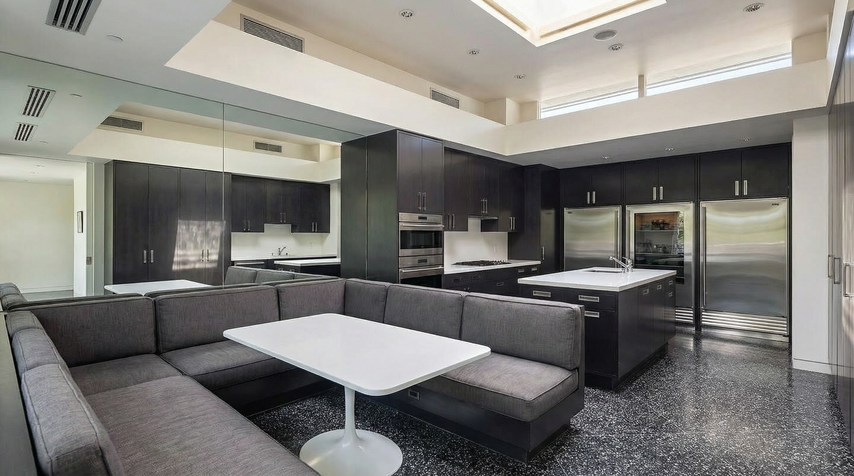 This interior shot displays a high-end kitchen featuring dark cabinetry, stainless steel appliances, and a kitchen island topped with white countertop. A unique built-in booth seating with a white table complements the kitchen area, along with the polished terrazzo flooring. The design incorporates modern elements and a spacious layout, creating a stylish and functional space.