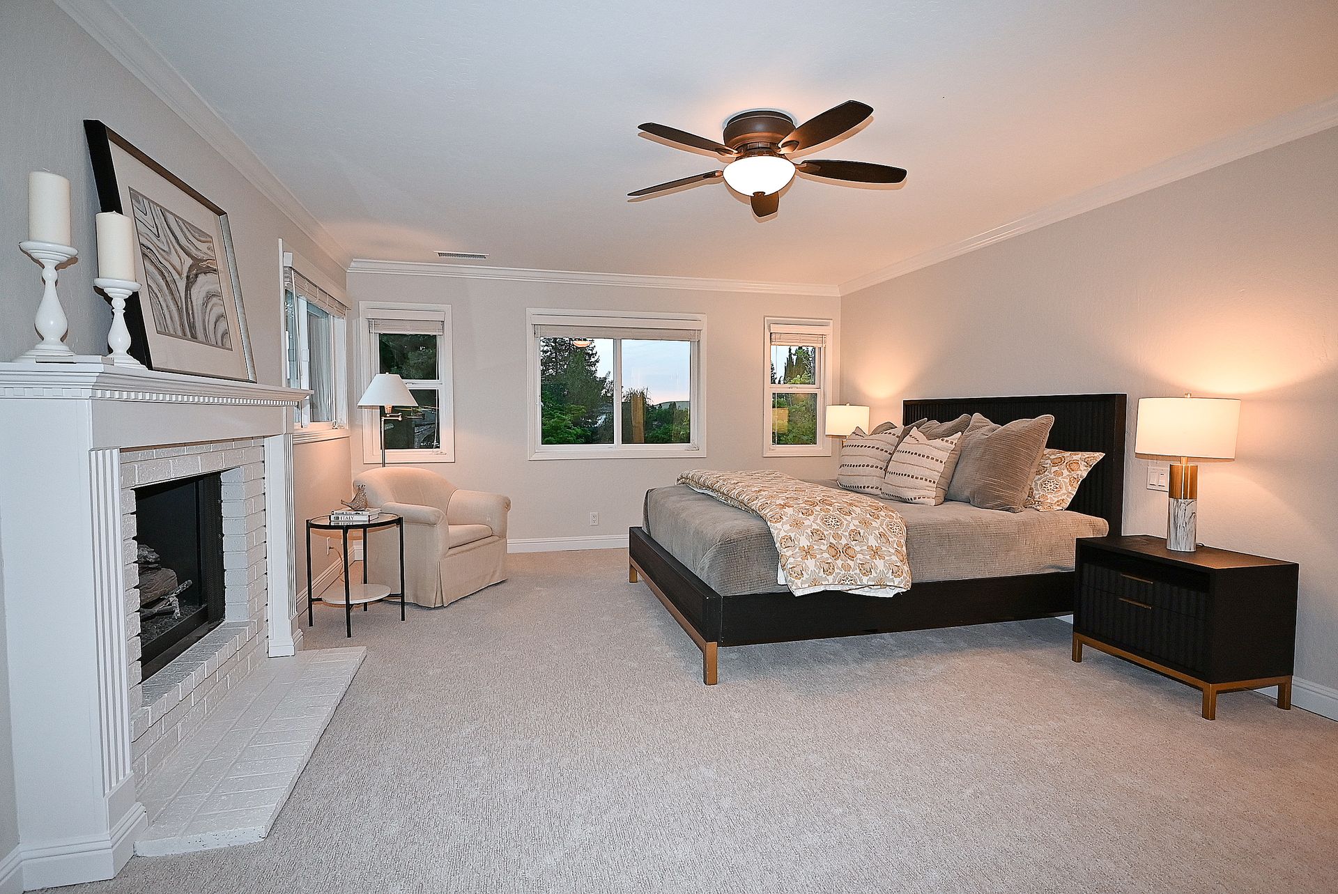This spacious primary bedroom features neutral carpeting, a large bed with modern bedding, and a cozy fireplace, creating an inviting and serene atmosphere. Large windows allow for ample natural light, while the clean, minimalist decor and warm lighting from the lamps and ceiling fan enhance the overall restful ambiance of the space.