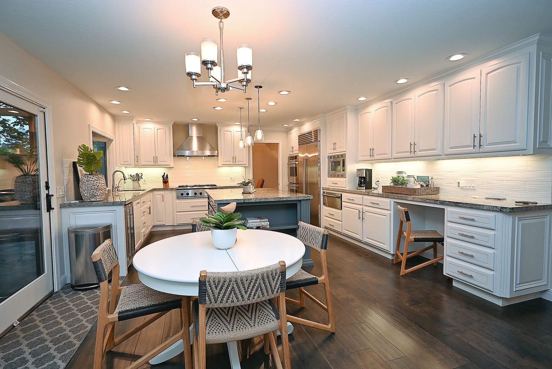 This bright and spacious kitchen features high-end white cabinetry, dark granite countertops, and professional-grade stainless steel appliances. The layout includes a central island, a built-in desk workstation, and an adjoining dining area with woven chairs, creating a functional yet elegant open-concept design. Warm interior lighting combined with recessed ceiling lights and a stylish chandelier provides a welcoming and upscale atmosphere.
