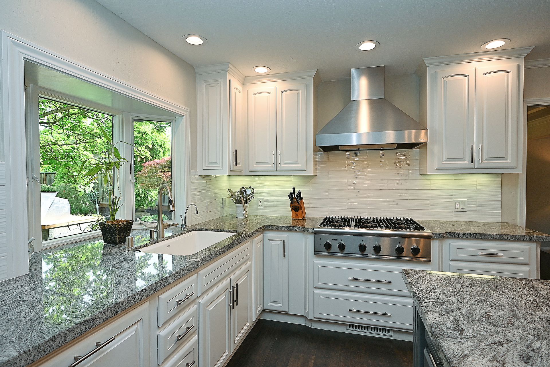 This bright, well-appointed kitchen features classic white Shaker-style cabinetry contrasted by speckled granite countertops and a clean, horizontal-tiled backsplash. A stainless steel gas range with a matching range hood serves as the focal point, while the adjacent sink area looks out onto lush greenery through a large, corner-wrapped window. The space feels refined and functional, with an open layout that incorporates modern appliances and dark hardwood flooring.