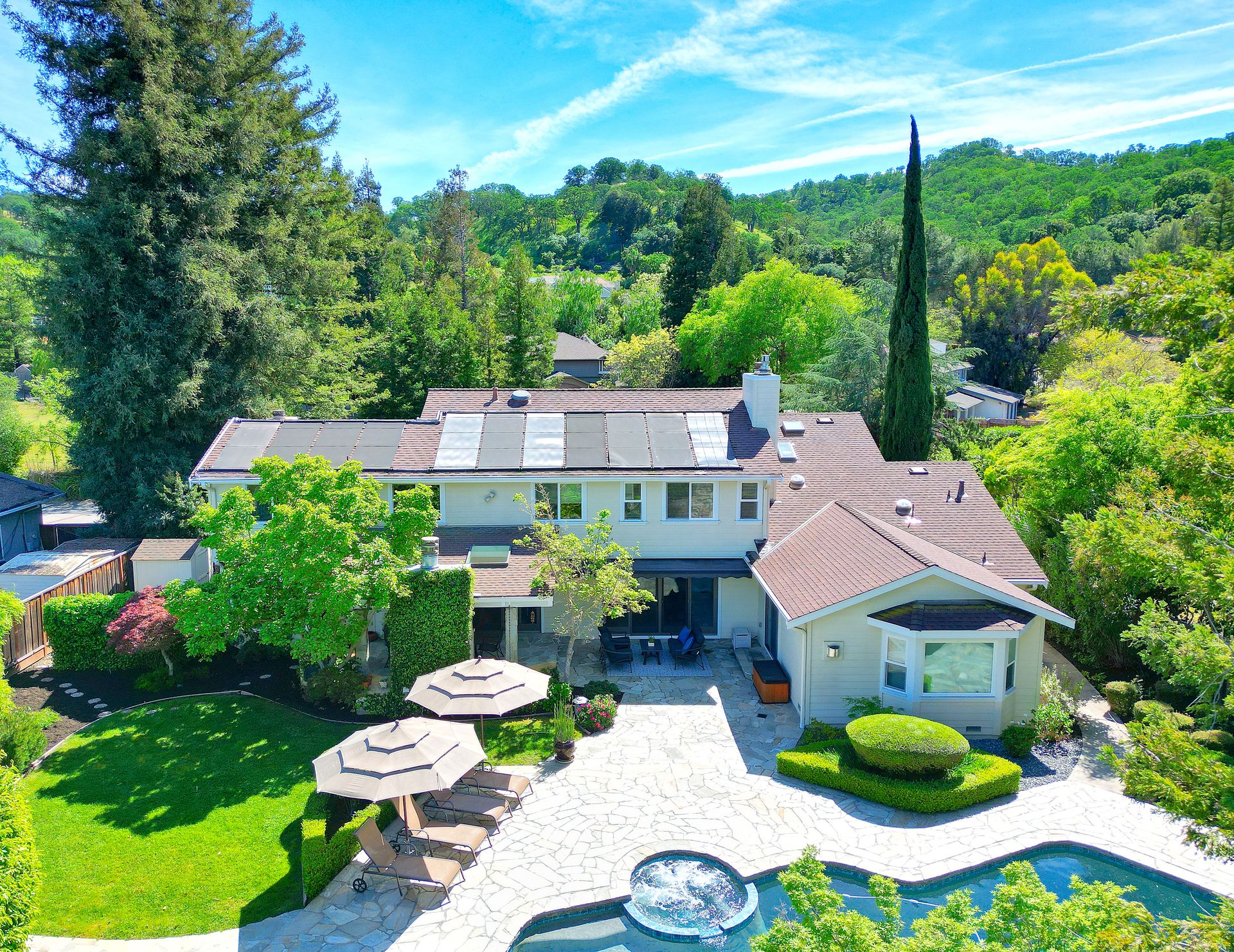 This stunning aerial view highlights a spacious suburban residence, featuring a large backyard complete with a stone-paved patio, an inviting swimming pool, a spa, and lounge chairs. The property showcases meticulous landscaping including manicured shrubs and shade trees, contrasted by a prominent roof equipped with solar panels. The overall impression is one of luxury, comfort, and an ideal setting for outdoor entertaining.