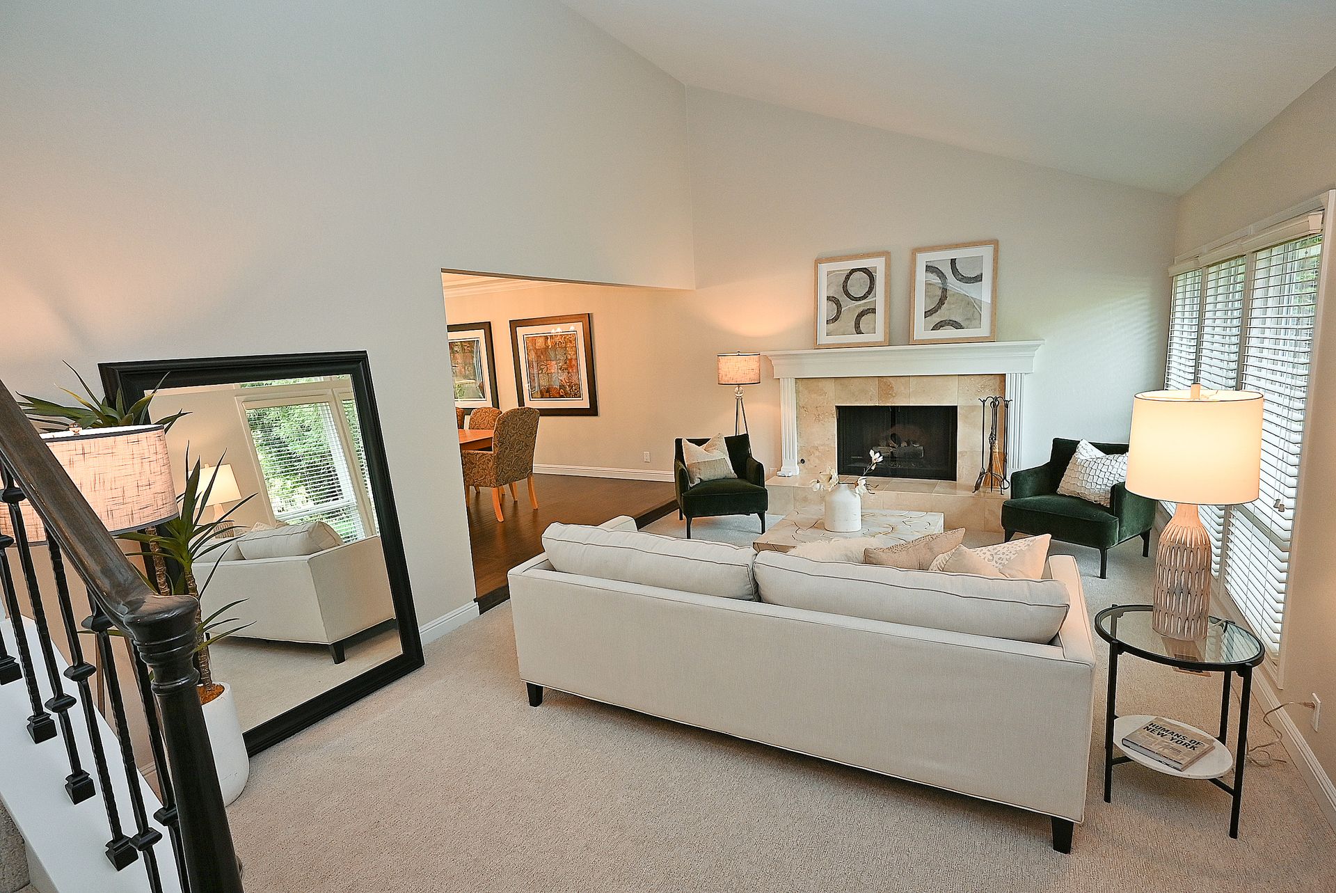 This bright and inviting living room features high vaulted ceilings, a cozy fireplace with a classic mantel, and plush neutral-toned furniture. A large floor mirror acts as a stylish focal point, reflecting the airy space and adding depth to the room's open-concept layout. The transition to the adjacent dining area, accented with warm wood floors and elegant art, creates a cohesive and comfortable flow throughout the home.