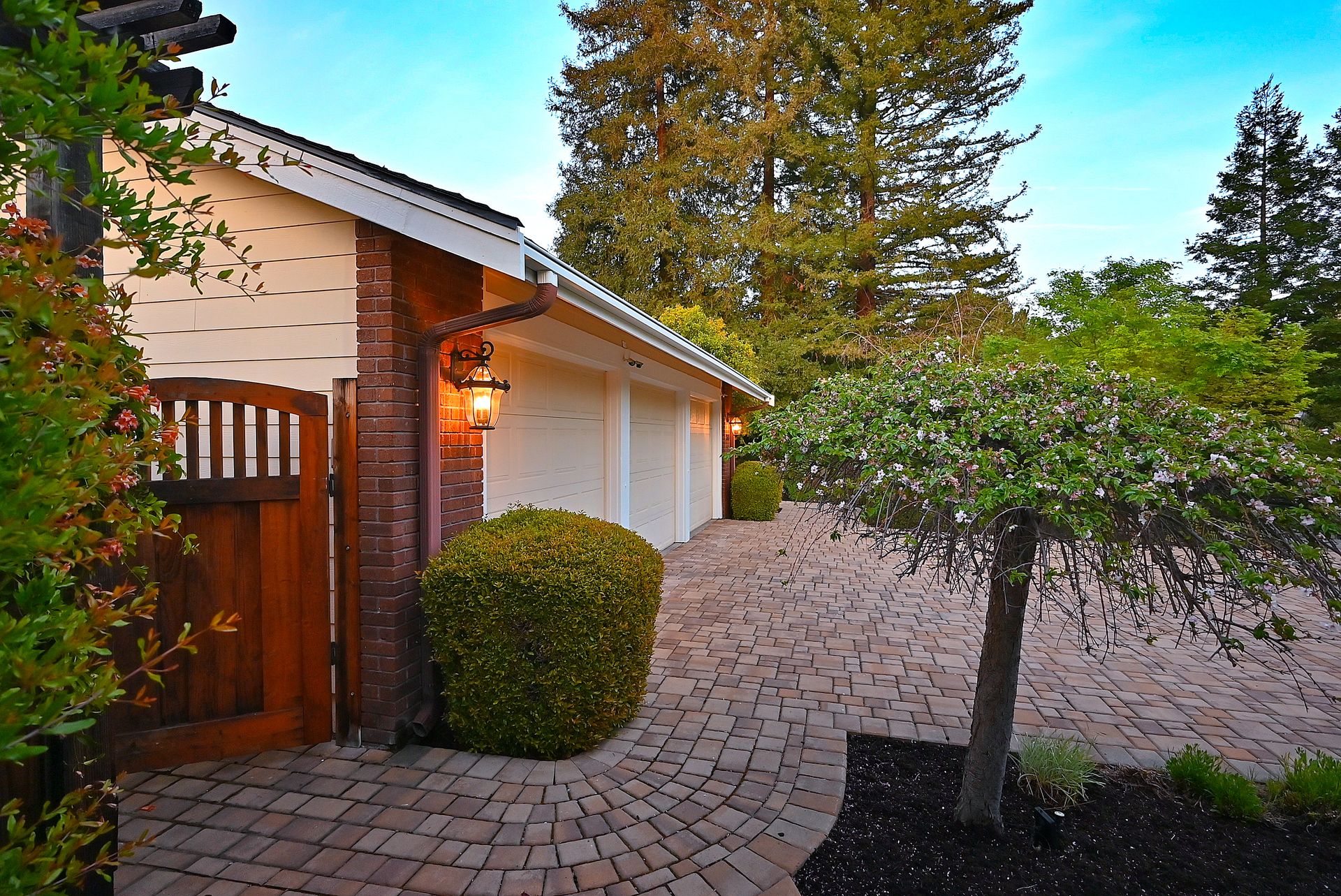 This well-maintained property features a three-car garage with light-colored siding and an attractive brick pillar accent. A curved paver driveway complements the lush landscaping, which includes a mature weeping cherry tree and manicured shrubs. The presence of warm wall-mounted lanterns creates an inviting aesthetic perfect for curb appeal.