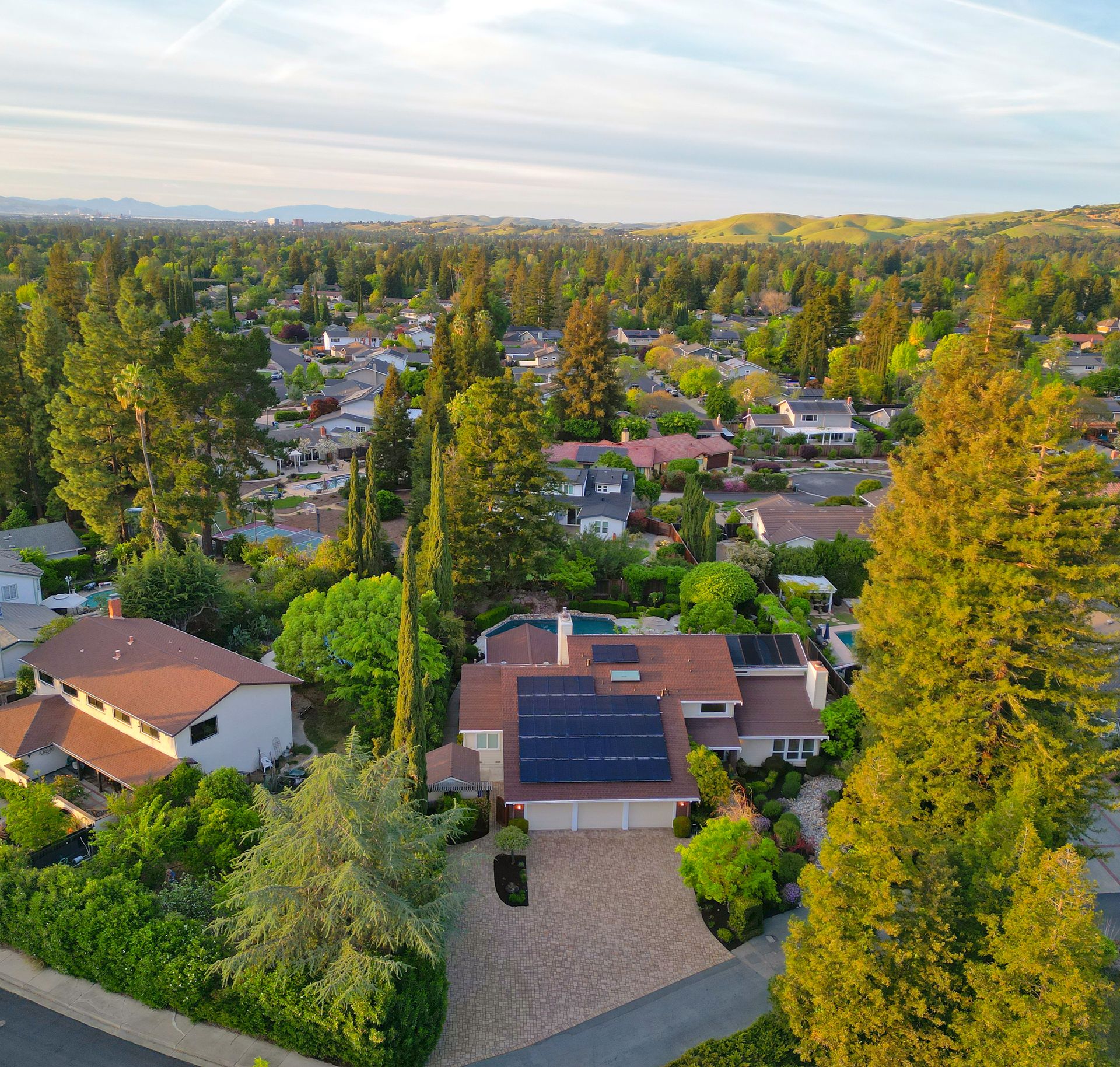 This is an aerial view showcasing a suburban residential property nestled in a lush, tree-filled landscape. The home features a prominent roof with an extensive solar panel array, a paver driveway, and a private swimming pool in the backyard. The setting is serene and verdant, with views of rolling hills in the background, suggesting a quiet, established neighborhood.