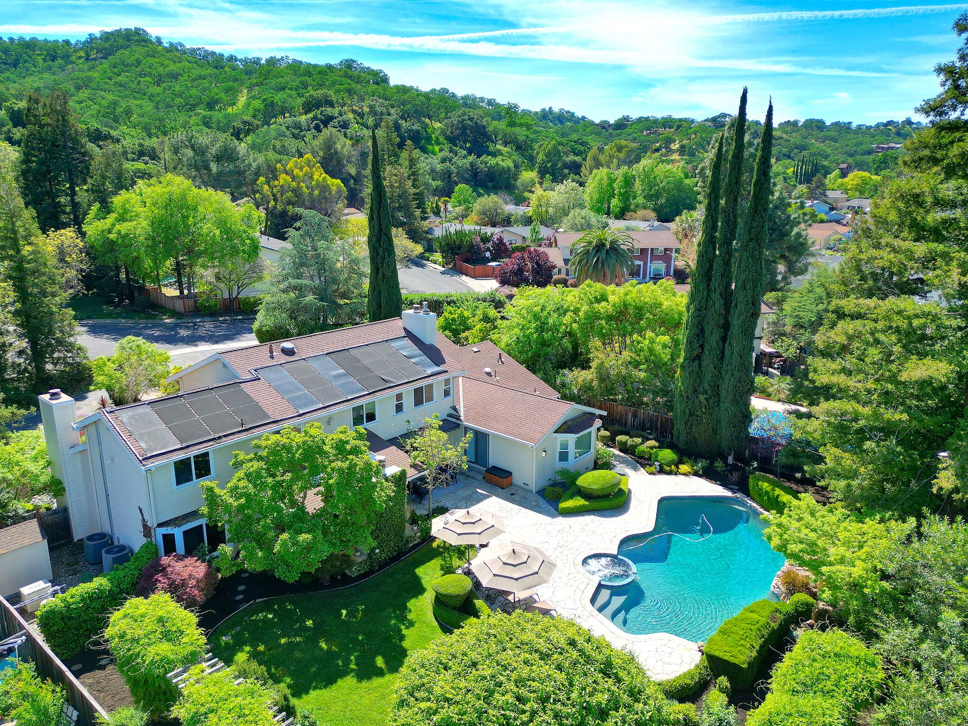 This stunning aerial view highlights a spacious suburban residence with a well-maintained backyard featuring a large swimming pool, spa, and stone patio area with umbrellas for relaxing. The home, which includes solar panels on a multi-pitched roof, is surrounded by lush green landscaping, mature trees, and rolling hills in the background. It offers a perfect blend of privacy and outdoor living, ideal for entertaining and family recreation.