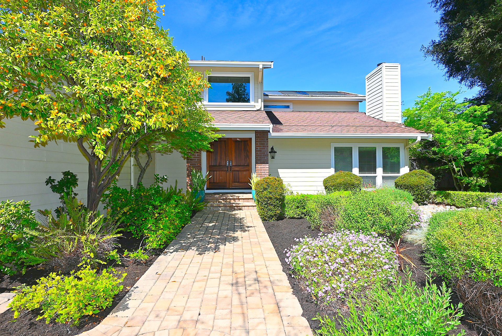 This charming front view showcases a well-maintained home with a classic architectural style, featuring a prominent wood double-door entrance accessed by a clean paver walkway. The exterior is complemented by a lush garden, including a fruit-bearing tree and vibrant flowering shrubs, set against a bright blue sky. A paved path leads directly to the front entrance, highlighting the inviting and manicured curb appeal of the property.