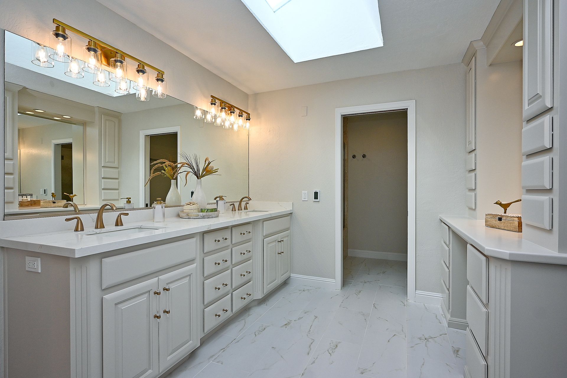 This elegant primary bathroom features double vanities with white shaker cabinets, gold-tone hardware, and pristine marble-patterned countertops. The space is brightened by a large skylight and sophisticated vanity lighting, while the monochromatic color palette and clean lines create a luxurious, modern feel.