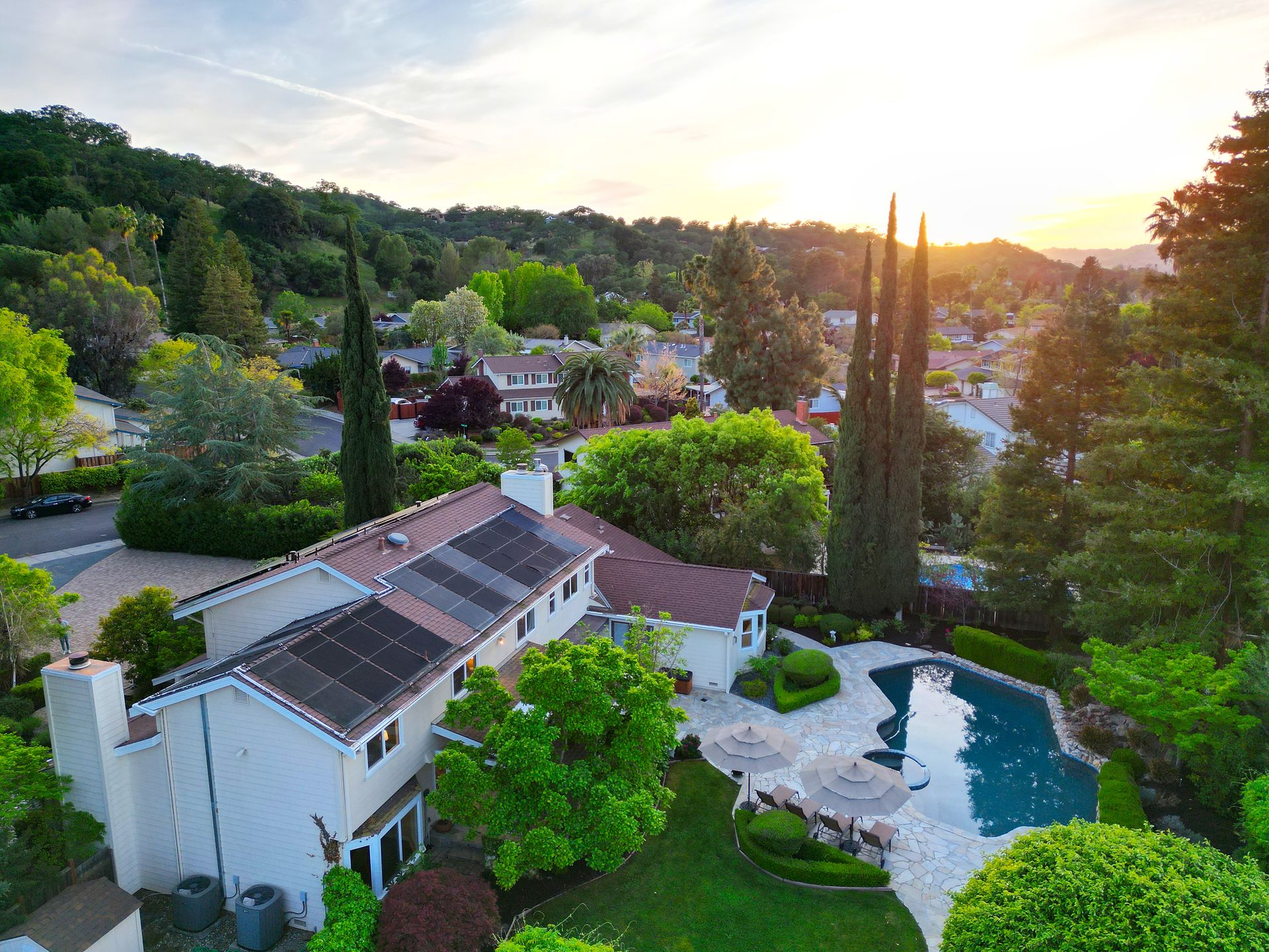 This high-angle, aerial perspective captures a spacious residential property featuring a large backyard with a private swimming pool, flagstone patio, and mature landscaping. The home itself boasts a multi-level design with a prominent solar panel installation on the roof, surrounded by trees and a scenic hillside neighborhood. The peaceful, sun-drenched setting suggests an appealing suburban lifestyle perfect for outdoor entertaining.