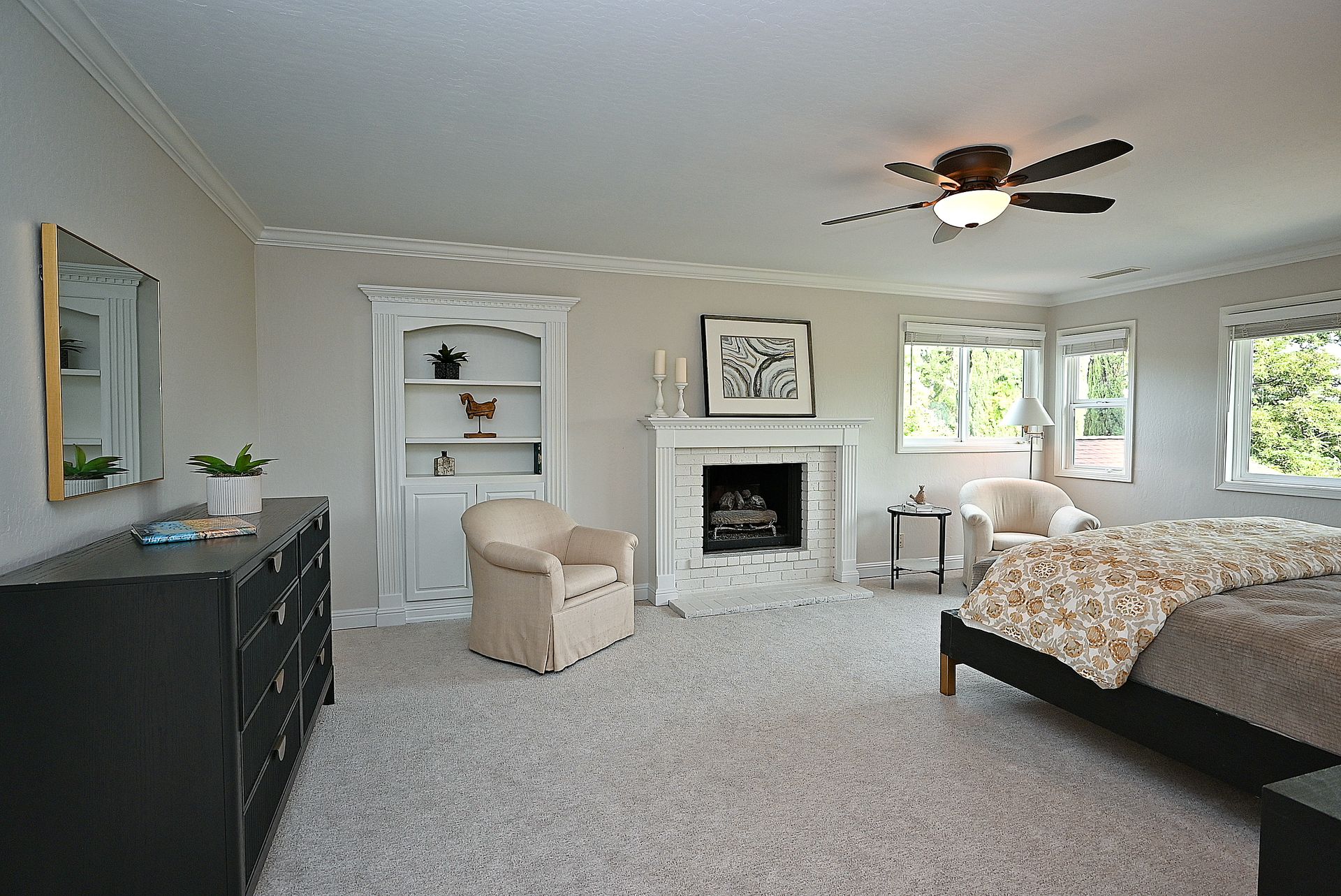This spacious primary bedroom features neutral wall tones, plush carpeting, and a cozy fireplace with an ivory brick surround. The room is furnished with a comfortable sitting chair, a dark dresser, and a large bed with a patterned comforter, creating a relaxing and inviting atmosphere. Plentiful natural light streams in through multiple windows, enhancing the airy and well-appointed feel of the space.