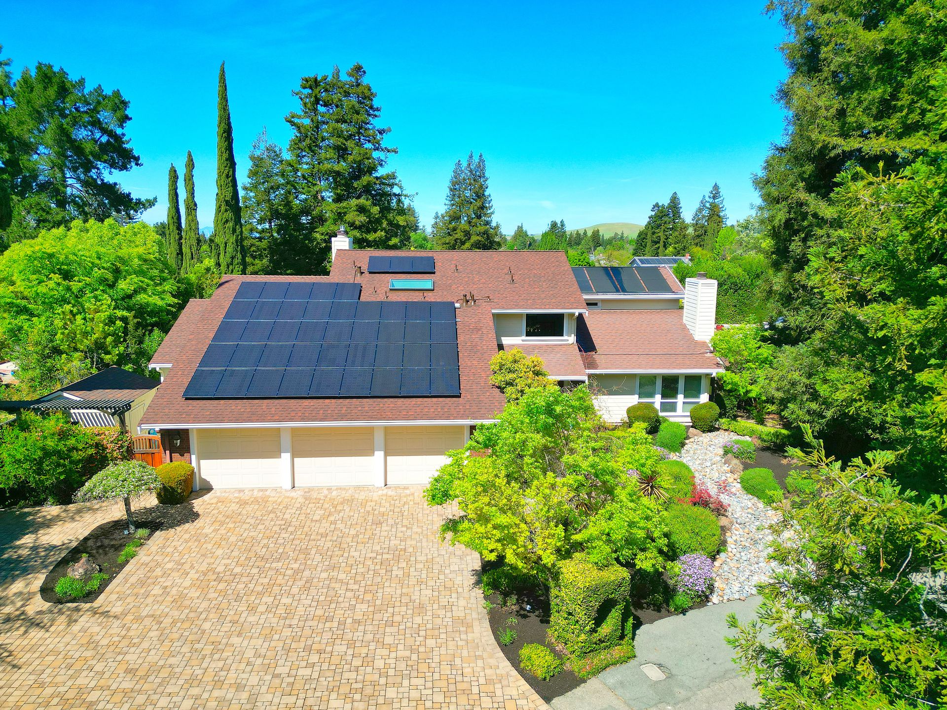 This is an aerial view of a suburban residence featuring a large driveway and a multi-car garage. The home is equipped with solar panels on the roof and is nestled within a lush, well-landscaped yard surrounded by mature trees. The property has a charming, well-maintained aesthetic with a brick-paved driveway that adds curb appeal.