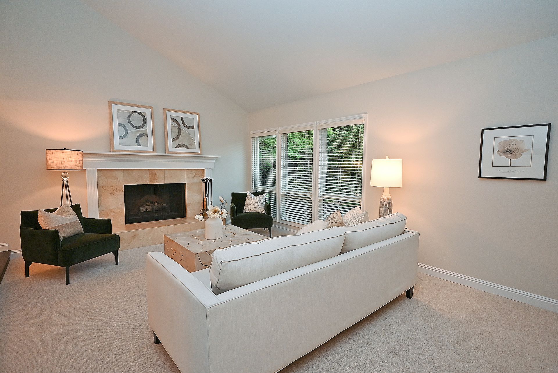 This bright and inviting living room features a vaulted ceiling, a spacious light-colored sofa, and two dark velvet accent chairs arranged around a textured square coffee table. The centerpiece is a stone fireplace flanked by elegant lamps and adorned with framed artwork, creating a cozy yet sophisticated atmosphere perfect for relaxation.