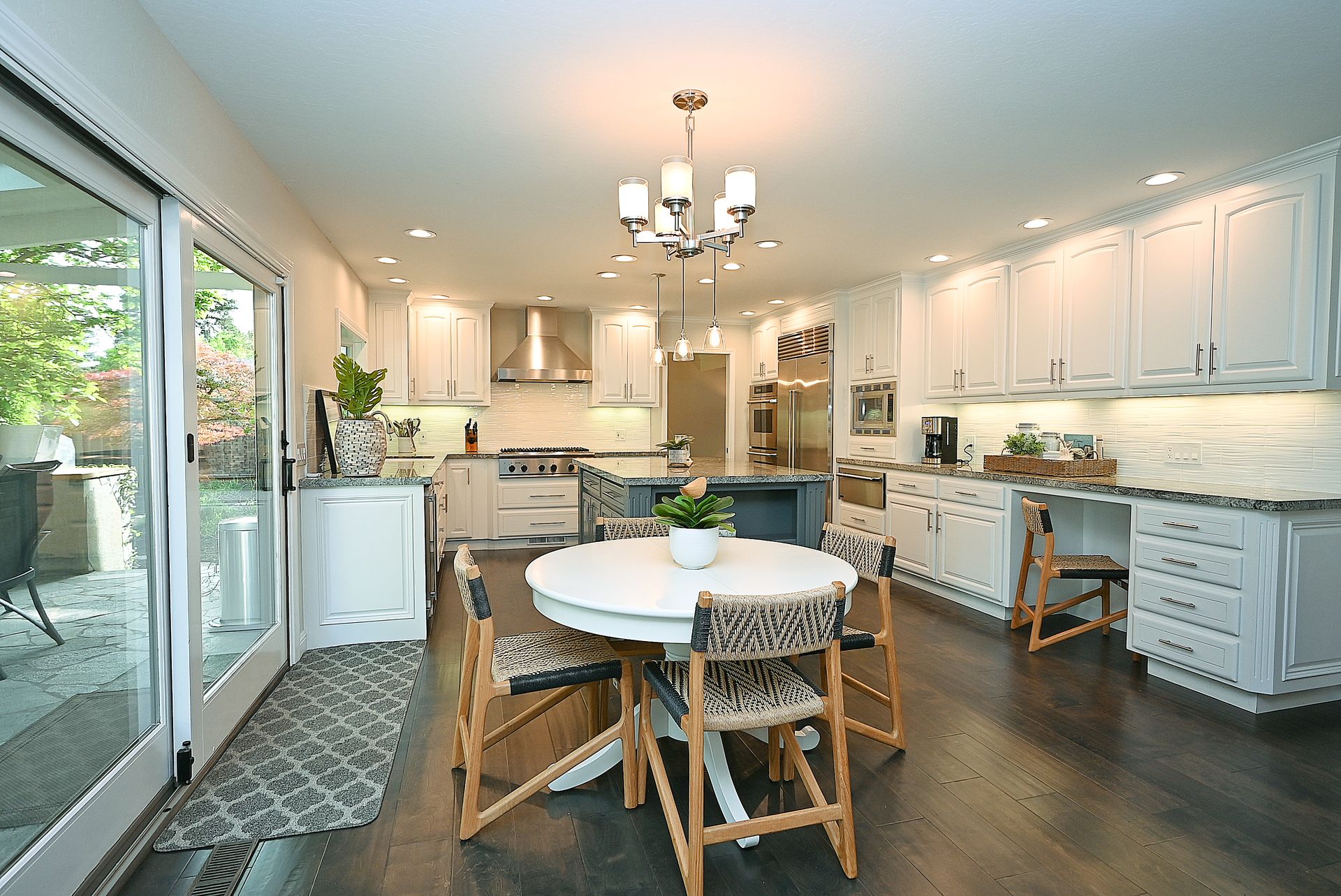 This bright and spacious kitchen features classic white cabinetry accented by a dark-toned central island, combining traditional elegance with modern functionality. It includes high-end stainless steel appliances, granite countertops, and a cozy dining area with a round table and woven chairs. The transition to the outdoor space through glass sliding doors, combined with warm hardwood flooring and ample recessed lighting, creates an inviting and airy atmosphere.