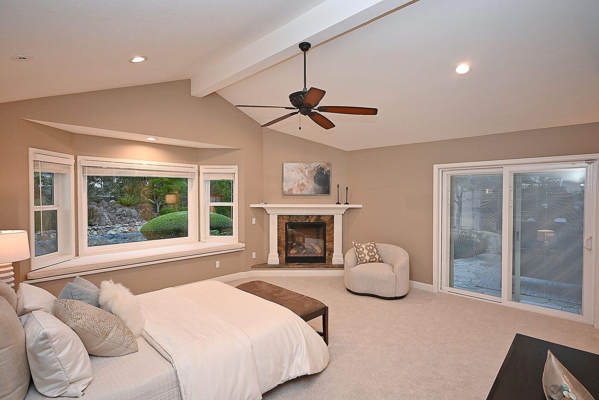 This spacious primary bedroom features neutral wall tones, a high vaulted ceiling with a wooden beam, and soft carpeting throughout. A cozy corner fireplace with a stone surround acts as a focal point, complemented by a large bay window seat looking out onto a landscaped backyard. The room is well-appointed with access to a private patio via sliding glass doors, creating a serene and comfortable retreat.