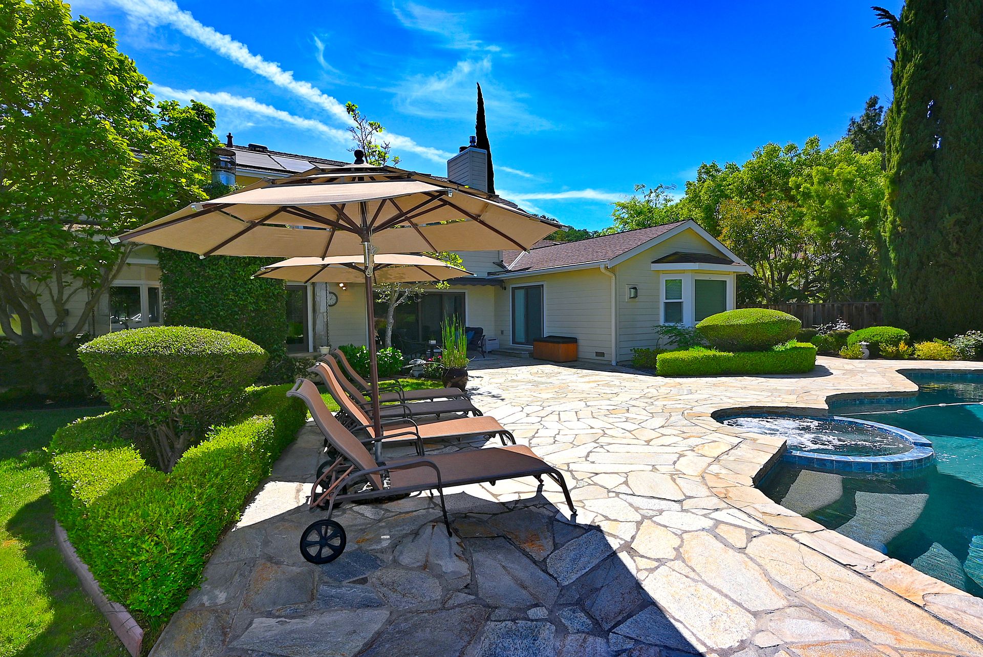 This inviting backyard features a stone-paved patio complete with a private pool and integrated spa. Multiple lounge chairs shaded by a large umbrella offer a relaxing seating area, complemented by manicured shrubbery and mature landscaping. The property offers a serene, resort-like atmosphere ideal for outdoor entertaining.