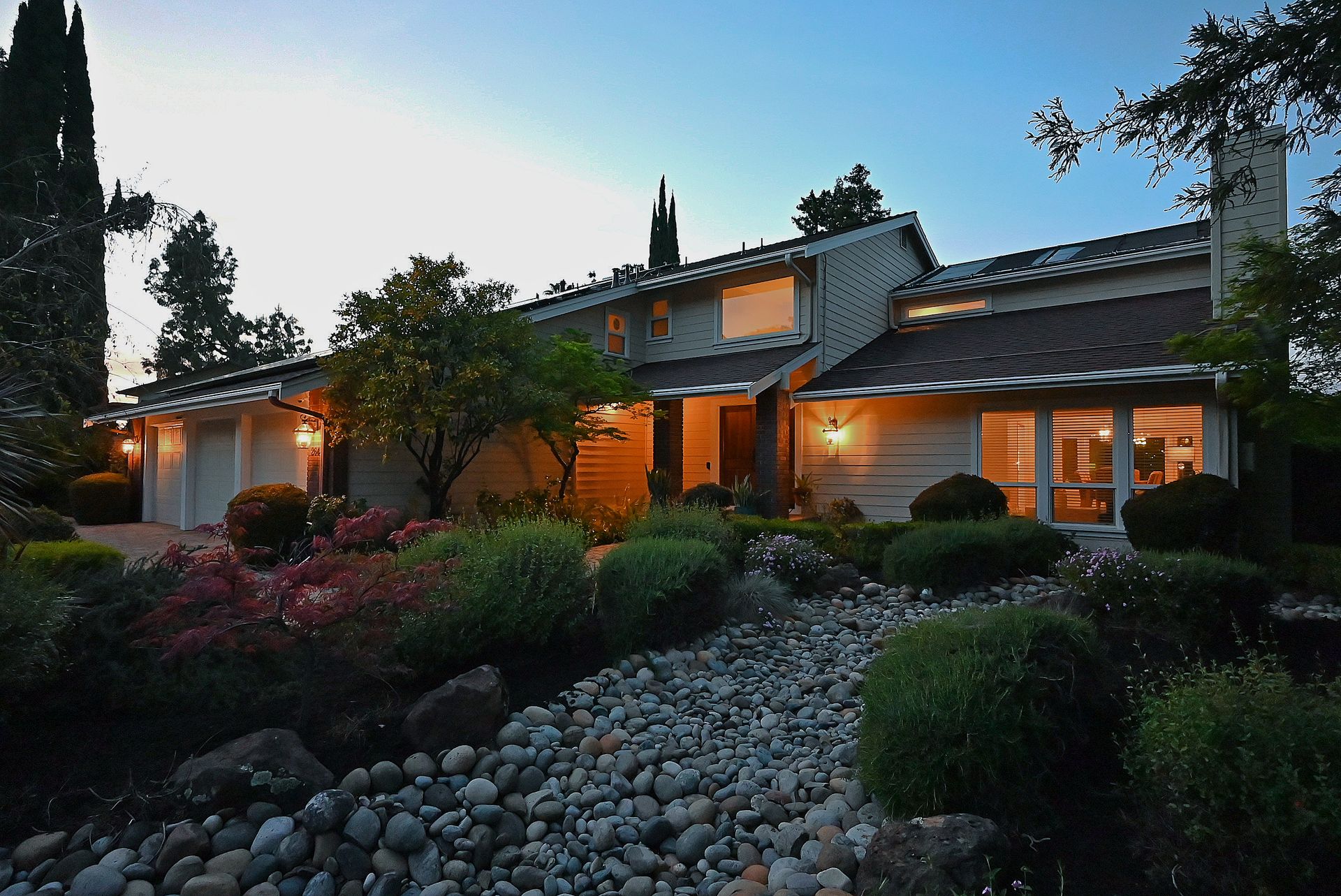 This charming, multi-level home features a well-manicured front yard with a unique dry-creek stone pathway leading to the main entrance. The exterior is accented with warm landscape lighting, showcasing the home's contemporary siding and large, inviting windows at twilight. The combination of established shrubbery, large trees, and the thoughtful rock garden creates a serene and polished curb appeal.