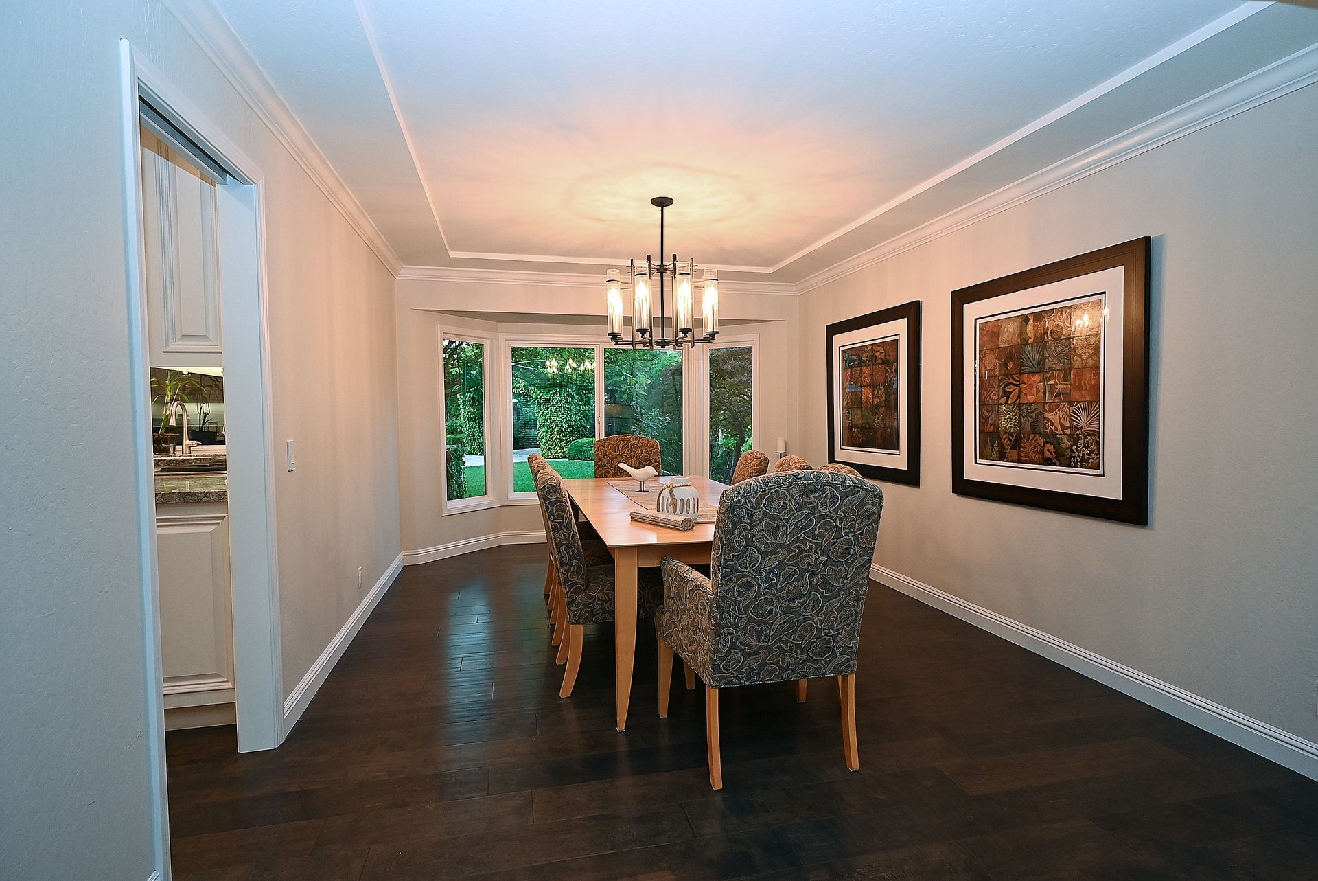 This elegant dining room features dark hardwood flooring and crown molding, centered around a sophisticated wooden dining table with patterned upholstered chairs. A modern multi-light chandelier acts as a focal point above the table, while the room is framed by a picture window offering a view of the outdoors and two large framed artworks on the side wall. The neutral color palette and clean lines create a refined atmosphere suitable for formal dining.