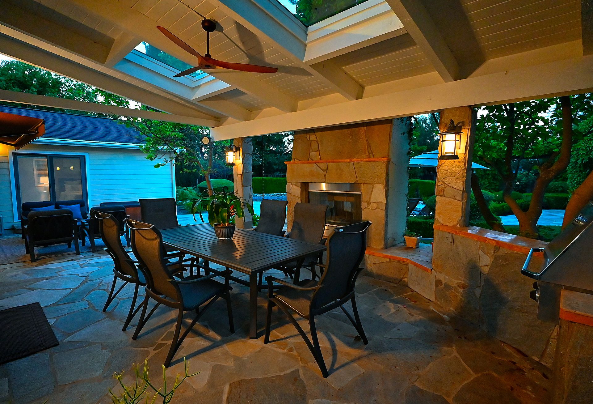 This inviting covered outdoor dining area features stone flooring, a built-in outdoor fireplace, and a dining set, making it perfect for entertaining. The space is topped with a rustic wooden ceiling, skylights, and a ceiling fan, all set against a backdrop of mature trees and a lush landscape. The warm lighting creates an intimate and welcoming atmosphere, highlighting the high-quality craftsmanship of the masonry.