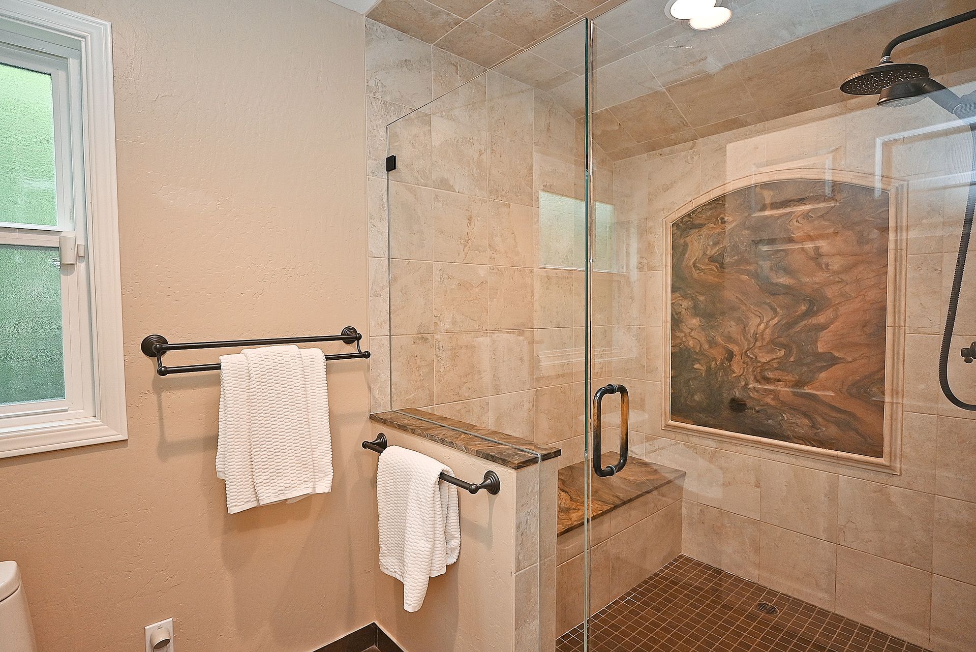 This primary bathroom features an upscale walk-in tile shower with a built-in stone bench, a decorative inset tiled backsplash, and a modern glass door assembly. The space is accented by bronze-finished plumbing fixtures, dual towel racks, and neutral tones that create a polished, spa-like atmosphere.