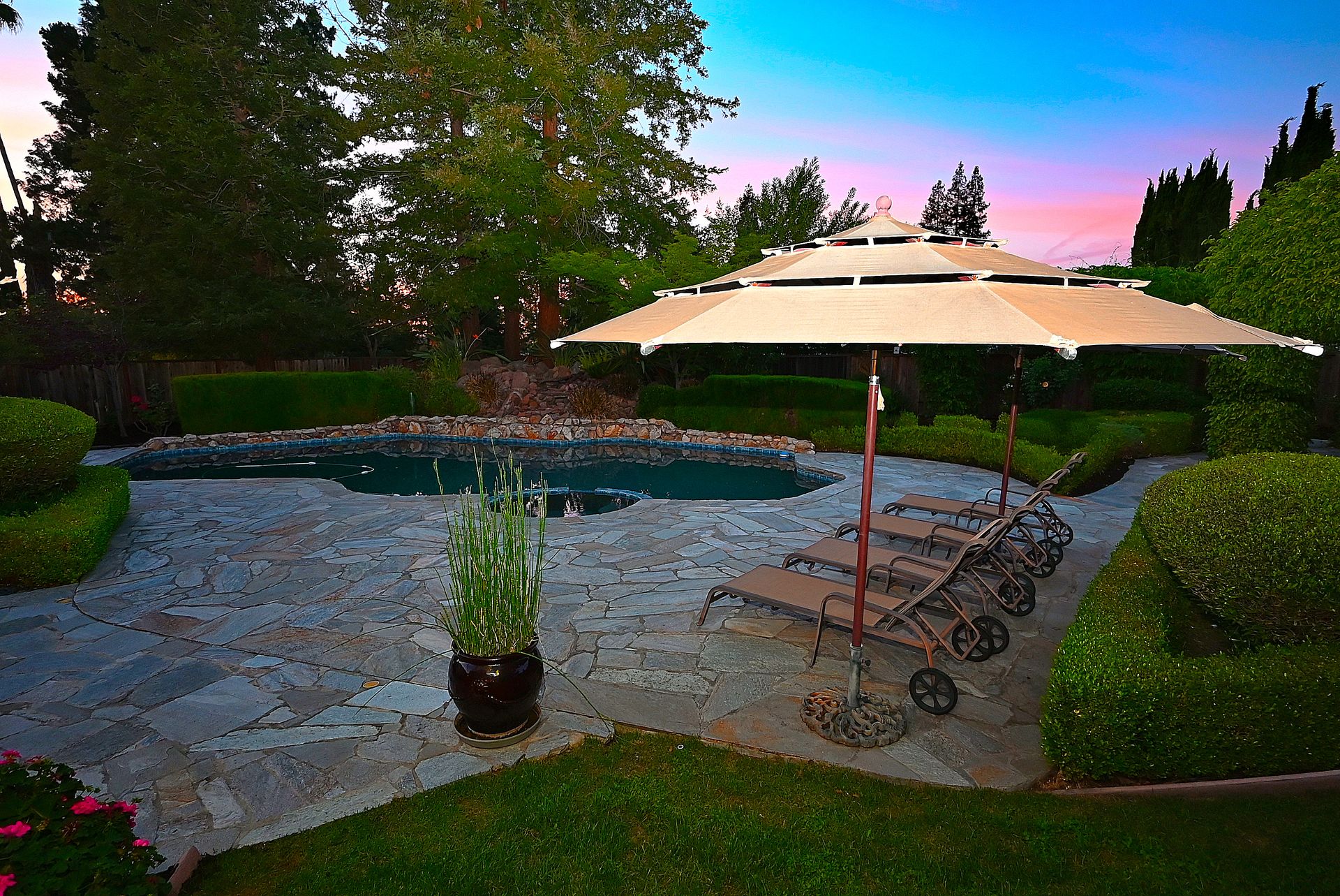 This stunning backyard oasis features a custom-shaped swimming pool integrated with a small spa, set against a backdrop of neatly manicured hedges and mature trees. The expansive flagstone patio provides plenty of space for outdoor leisure, centered around a large tan patio umbrella and multiple lounge chairs. The twilight lighting highlights the serene atmosphere, making it a perfect space for relaxing or entertaining.
