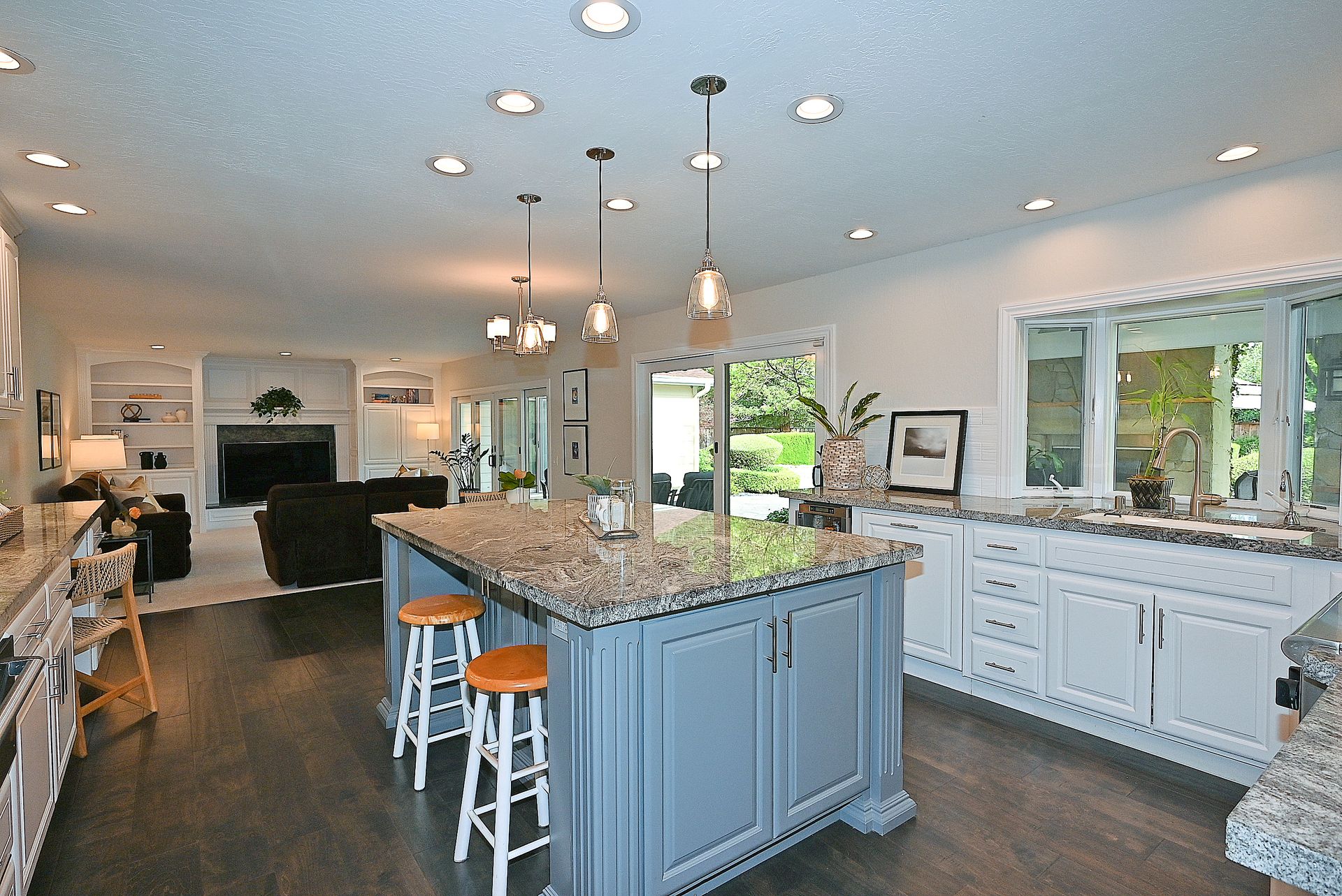 This spacious and well-lit kitchen features a large center island with a granite countertop, paired with a soft blue-grey cabinet finish that contrasts beautifully against the perimeter white cabinets. The open-concept layout flows seamlessly into a cozy living area with a fireplace, making it ideal for entertaining. High-end granite surfaces, elegant pendant lighting, and dark hardwood flooring create a sophisticated and inviting home environment.