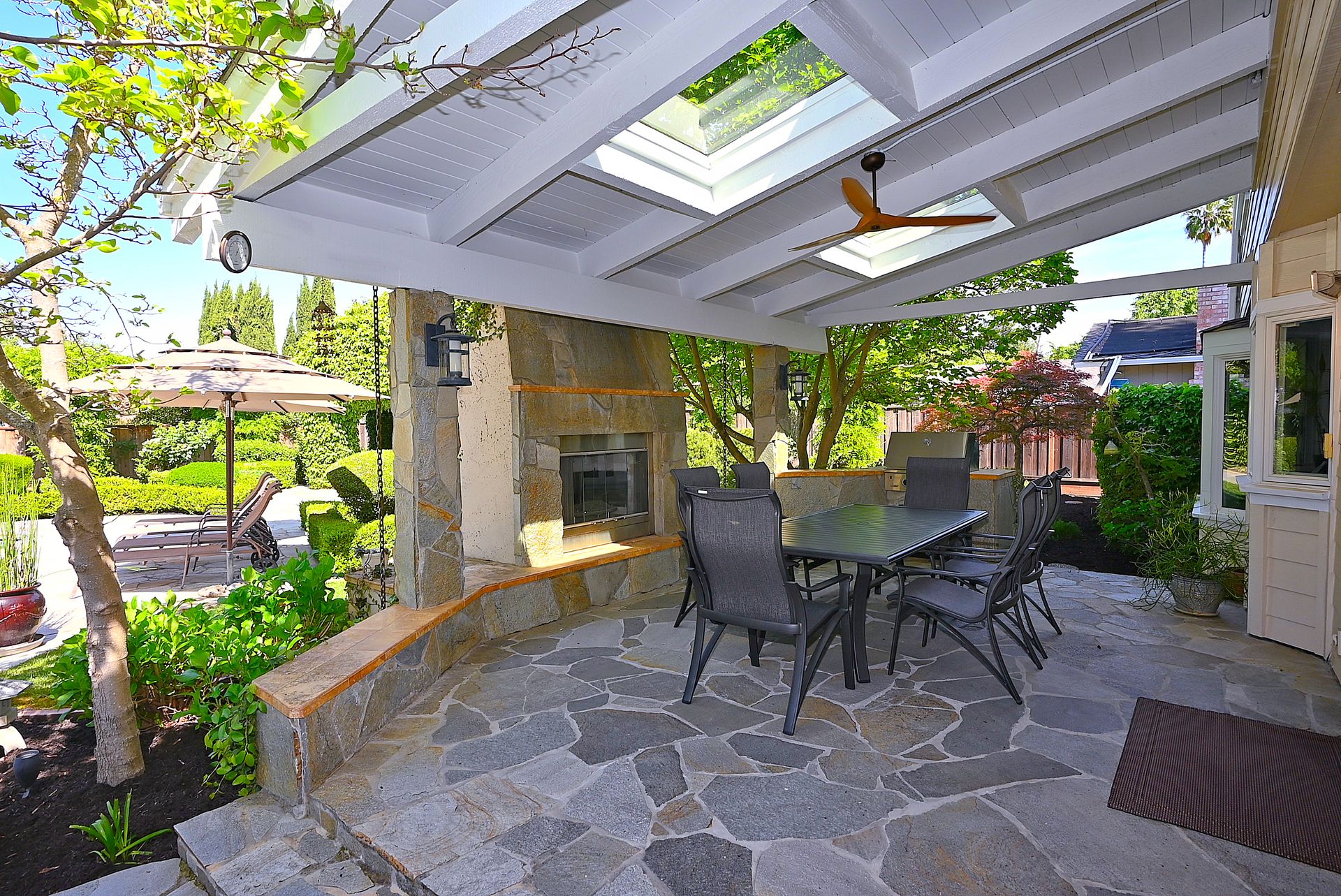 This spacious, covered outdoor patio features a stunning stone fireplace, flagstone flooring, and a dining set, making it an ideal space for entertaining. The area is sheltered by a white trellis roof with a skylight and a ceiling fan, seamlessly blending comfort and nature with views of a lush landscaped garden. This well-appointed outdoor living area adds significant value and appeal to a home's exterior amenities.