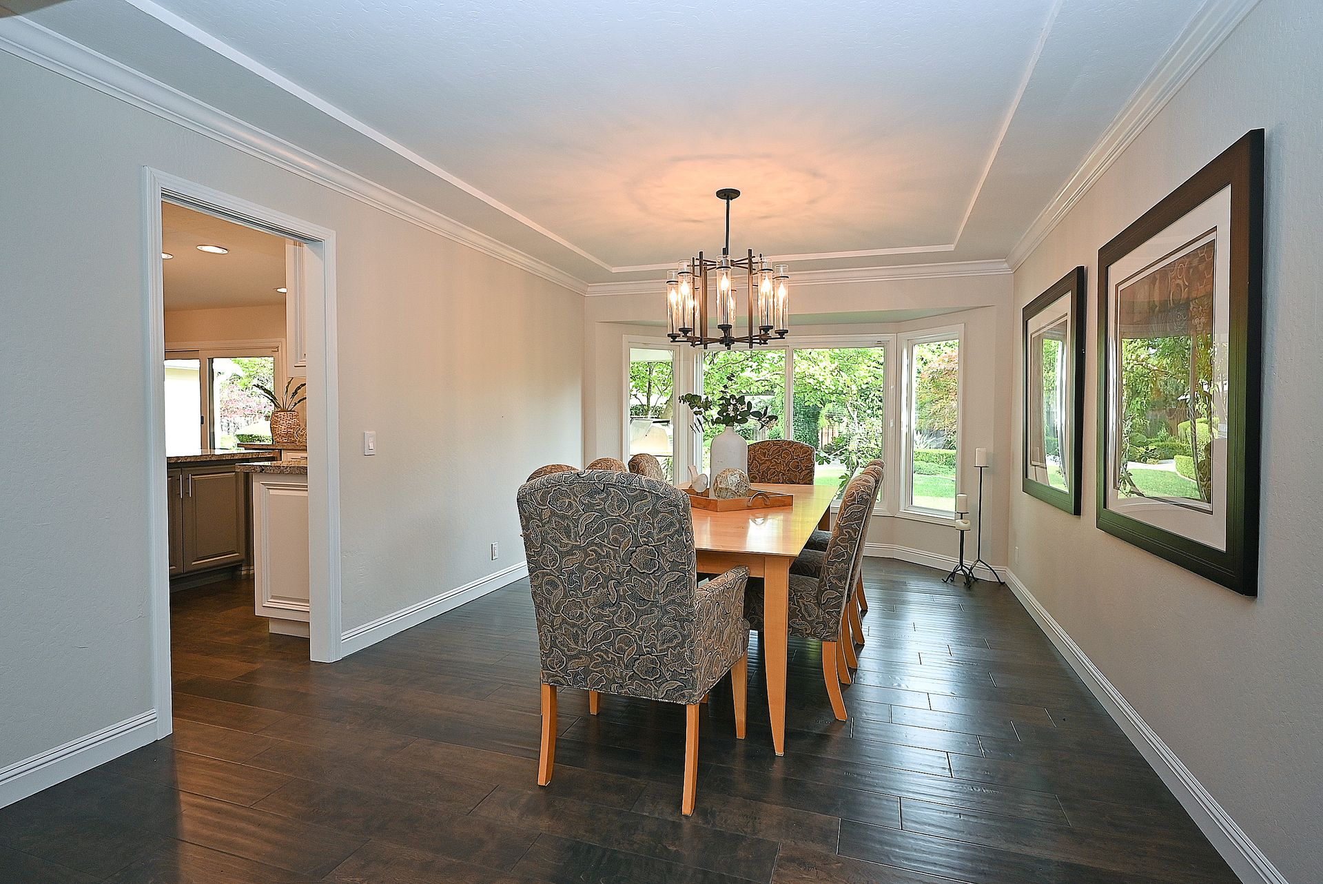 This elegant dining room features dark, wood-look flooring and a light-filled bay window that overlooks a lush garden. The space is anchored by a rectangular wood table with upholstered patterned chairs, topped by a sophisticated multi-light chandelier. Large framed art pieces on the wall create a formal, polished aesthetic suitable for both daily meals and gatherings.