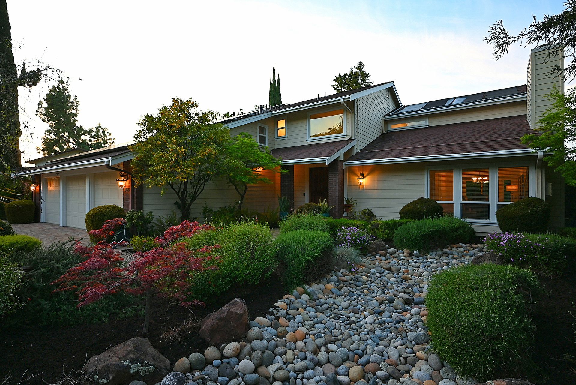 This charming, multi-level suburban home features an attractive landscape design with a stone-filled dry creek bed leading to the front entryway. The exterior showcases beige horizontal siding, a mix of gable rooflines, and multi-paned windows, complemented by warm lighting and a neatly paved driveway leading to the garage. The property exudes curb appeal with its manicured shrubs, a vibrant red Japanese maple, and thoughtful architectural details.