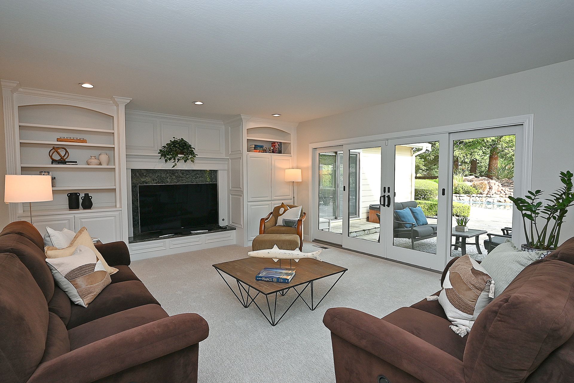 This inviting living room features plush brown seating, a uniquely designed wooden coffee table, and a large built-in fireplace and shelving unit that serves as the focal point. Large sliding glass doors provide a seamless transition to an outdoor patio, filling the room with natural light. The neutral color palette and clean lines create a cozy yet sophisticated atmosphere perfect for relaxing or entertaining.