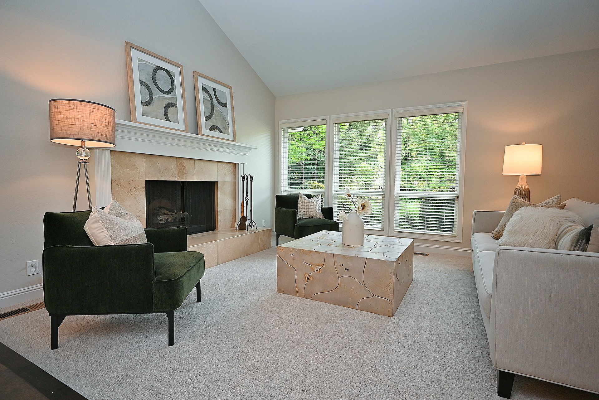 This bright and inviting living room features a vaulted ceiling and a tiled fireplace as the focal point, flanked by contemporary green armchairs. The room is anchored by a unique, light wood-textured coffee table and a soft neutral-toned sofa, creating a cozy and balanced conversation area. Large, sun-drenched windows with blinds look out onto lush greenery, enhancing the airy and relaxed aesthetic of the space.