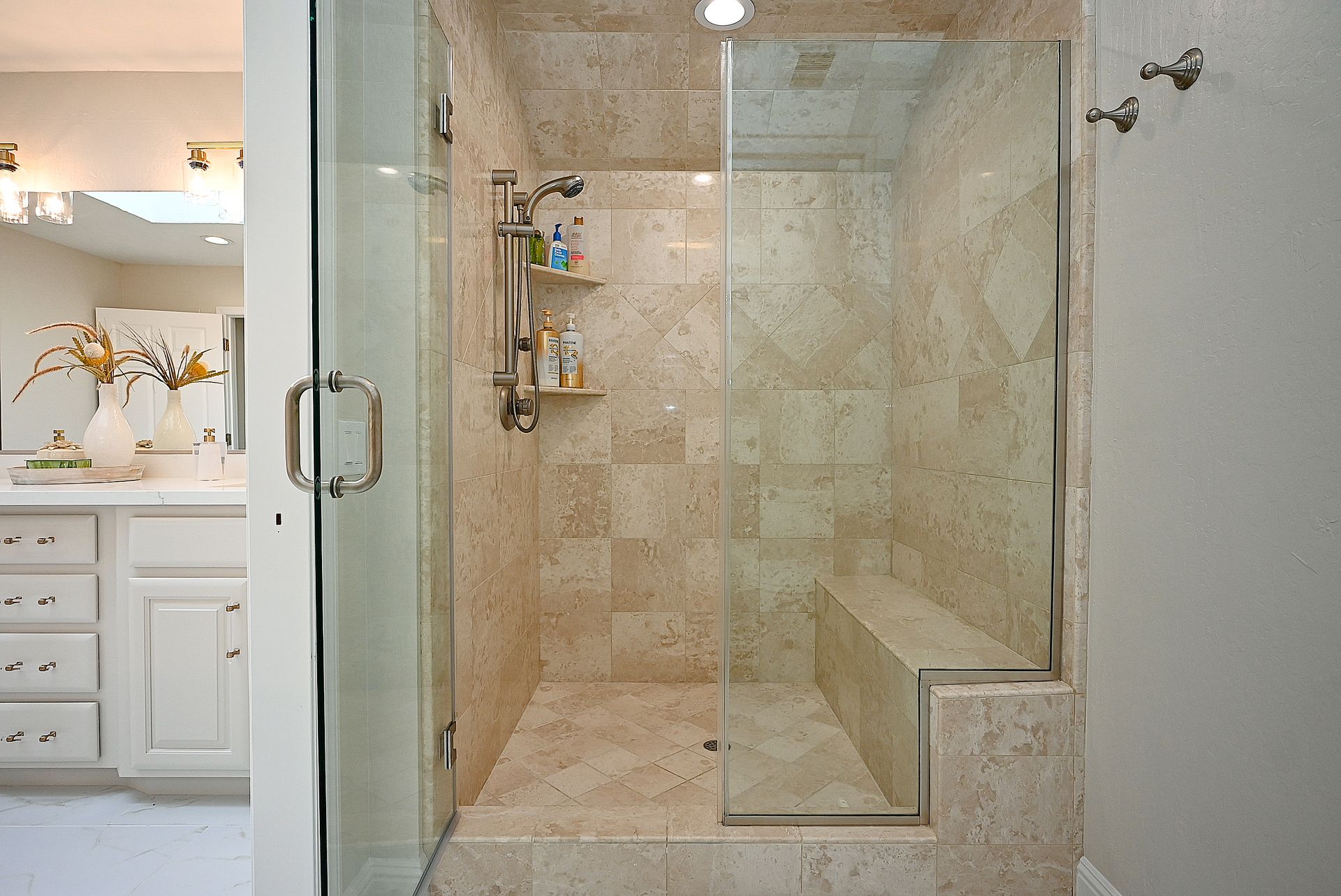 This walk-in shower features elegant, neutral-toned travertine tile throughout the walls and floor, complemented by a built-in bench and a glass enclosure. The shower is equipped with a modern nickel-finished rainfall showerhead and integrated storage shelves, reflecting a sophisticated and clean design. A glimpse of the adjacent vanity area suggests a cohesive and upscale primary bathroom aesthetic.