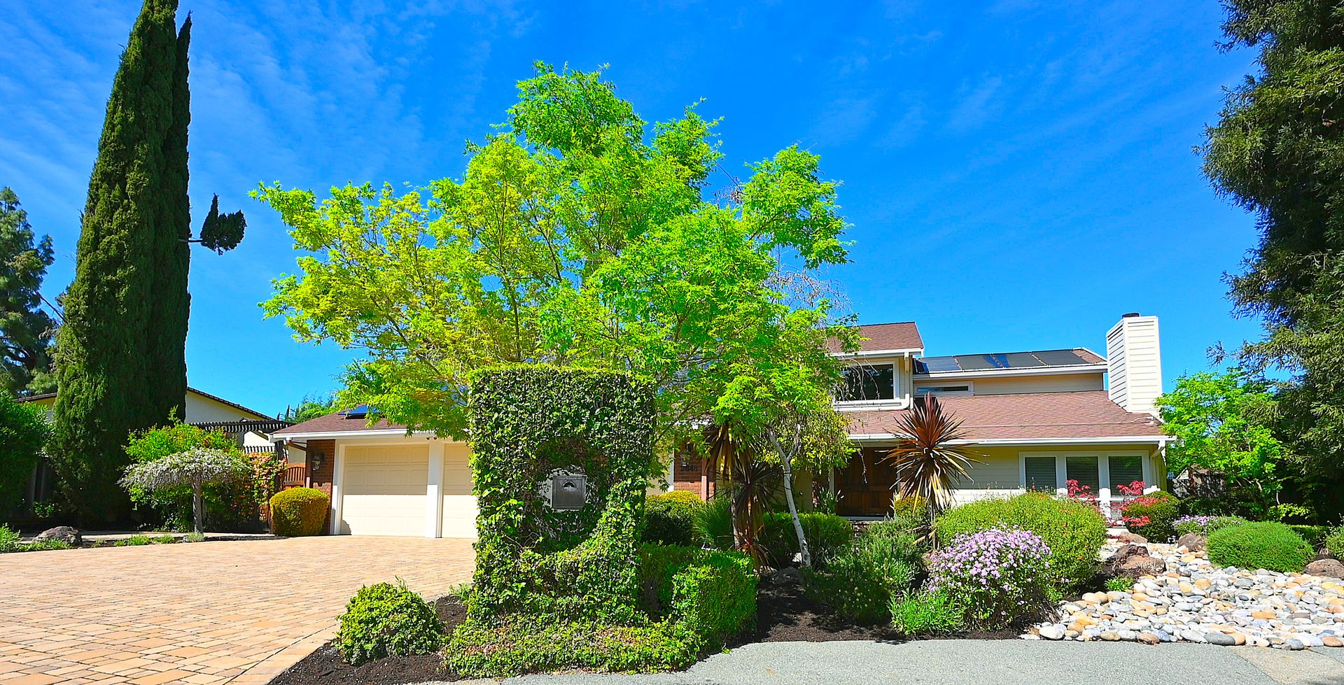 This charming suburban residence features a multi-level roofline with solar panels and a clean beige exterior. The property is highlighted by a beautifully landscaped front yard, including a distinct ivy-covered mailbox pillar, a mature tree, and cobblestone driveway accents. The home offers a welcoming curb appeal with a balanced mix of natural greenery and well-maintained hardscaping.