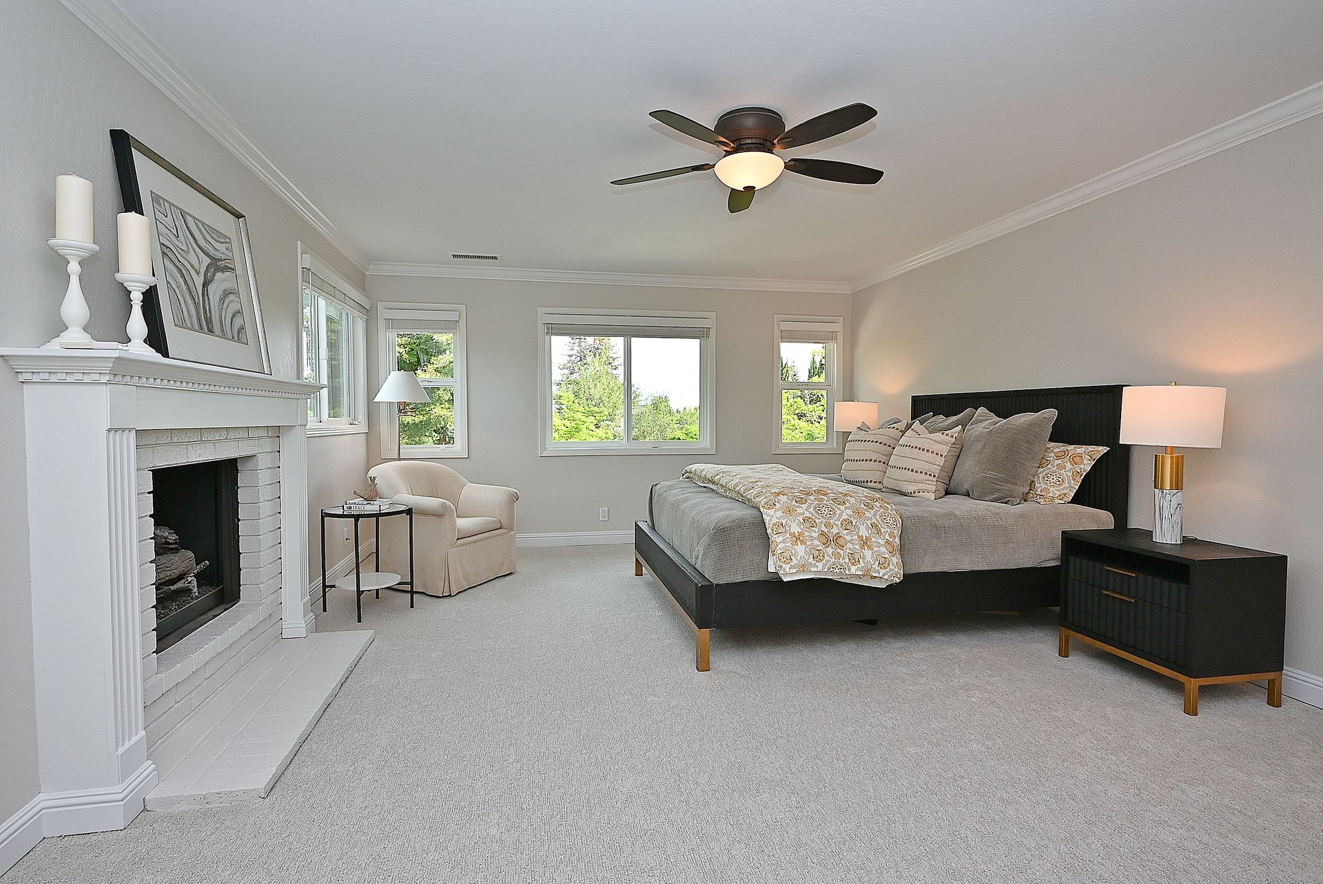 This spacious primary bedroom features a calming neutral color palette, plush carpeting, and a classic fireplace as a focal point. The room includes a large bed with a contemporary headboard, a cozy seating area by the window, and stylish bedside tables with modern lamps. Warm natural light fills the space, creating a serene and inviting atmosphere.
