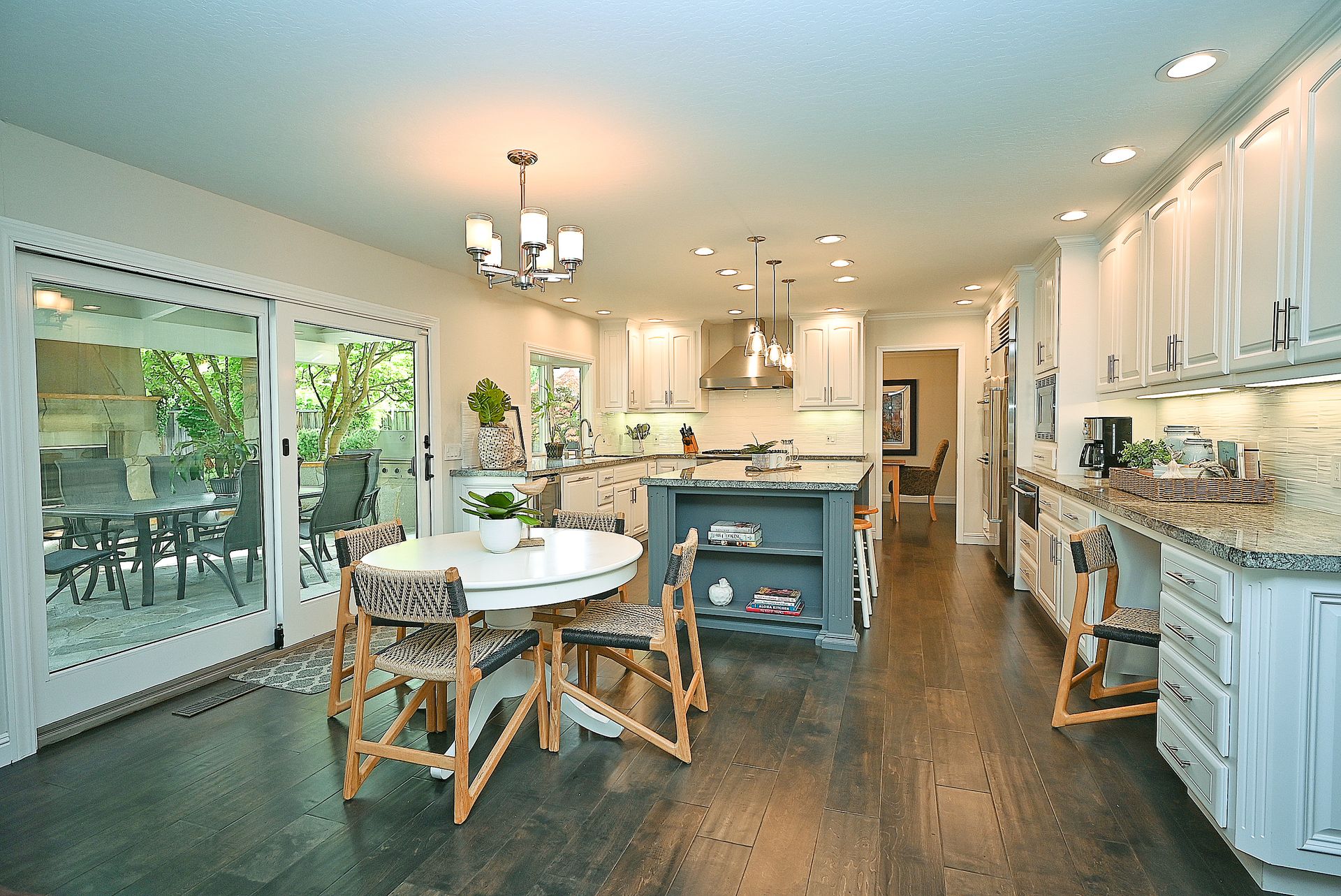 This spacious, modern kitchen features white cabinetry paired with a distinct dark gray kitchen island, creating a sophisticated contrast. The open-concept design flows seamlessly into a bright dining area with glass sliding doors that open to a patio, highlighting an airy and well-lit atmosphere. With its dark wood flooring, high-end finishes, and integrated seating at the island and counter, this room is both functional and aesthetically inviting.