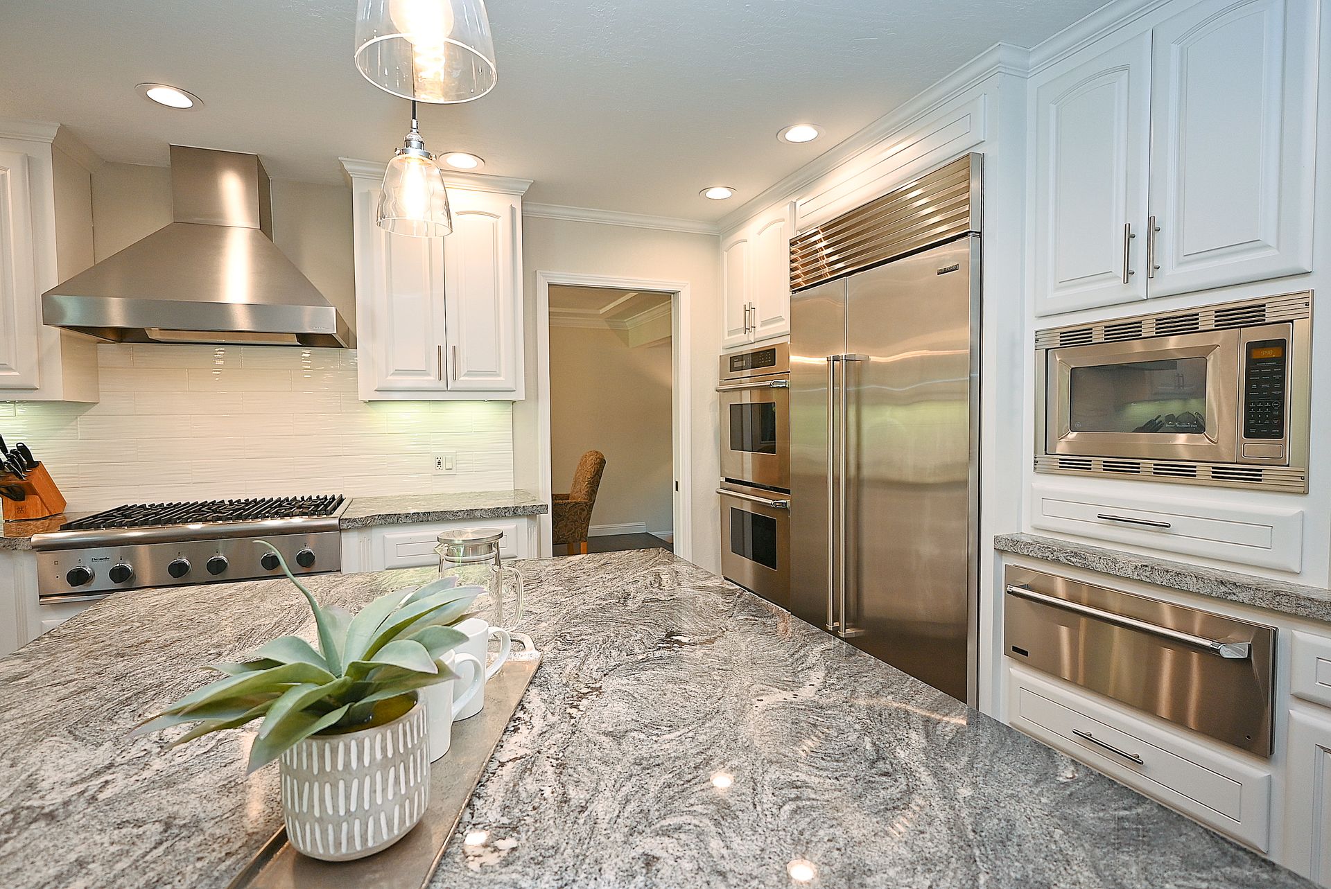 This kitchen features a sleek, modern design with custom white cabinetry, high-end stainless steel appliances, and premium granite countertops. The spacious island, topped with striking patterned stone, serves as the centerpiece, complemented by elegant pendant lighting and a professional-grade range. The space balances sophisticated function and style, making it an ideal environment for cooking and entertaining.