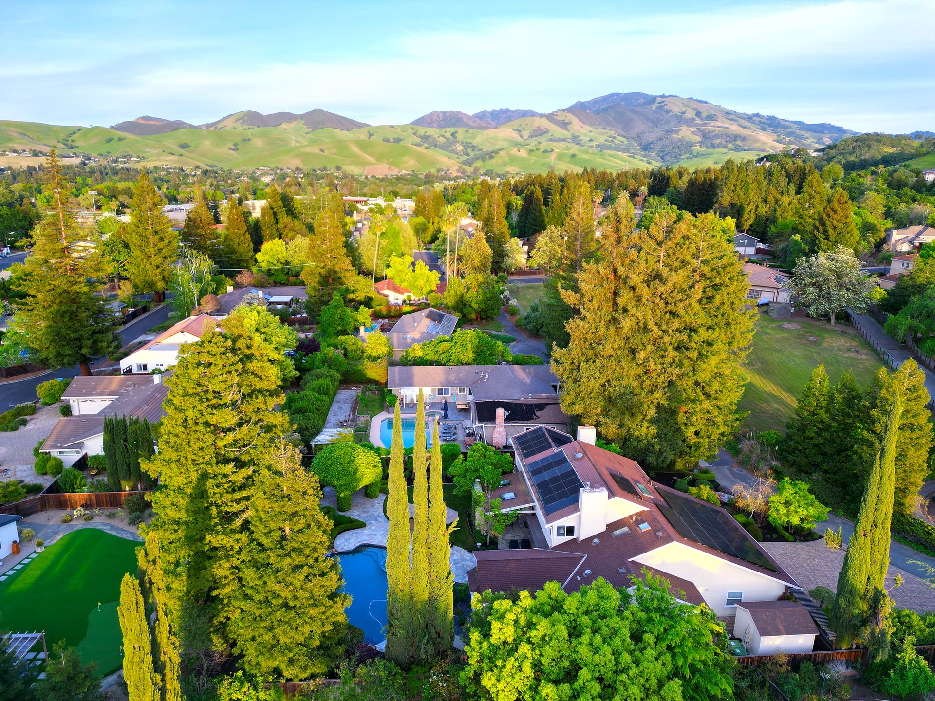 This high-angle, drone-style aerial photograph captures a luxurious suburban residence nestled against a scenic backdrop of rolling, sun-drenched hills. The property features a prime layout with expansive green lawns, a sparkling swimming pool, manicured gardens, and solar panel installations on the roof. The integration of lush mature trees and well-maintained landscaping provides a serene, private atmosphere in a highly desirable neighborhood setting.