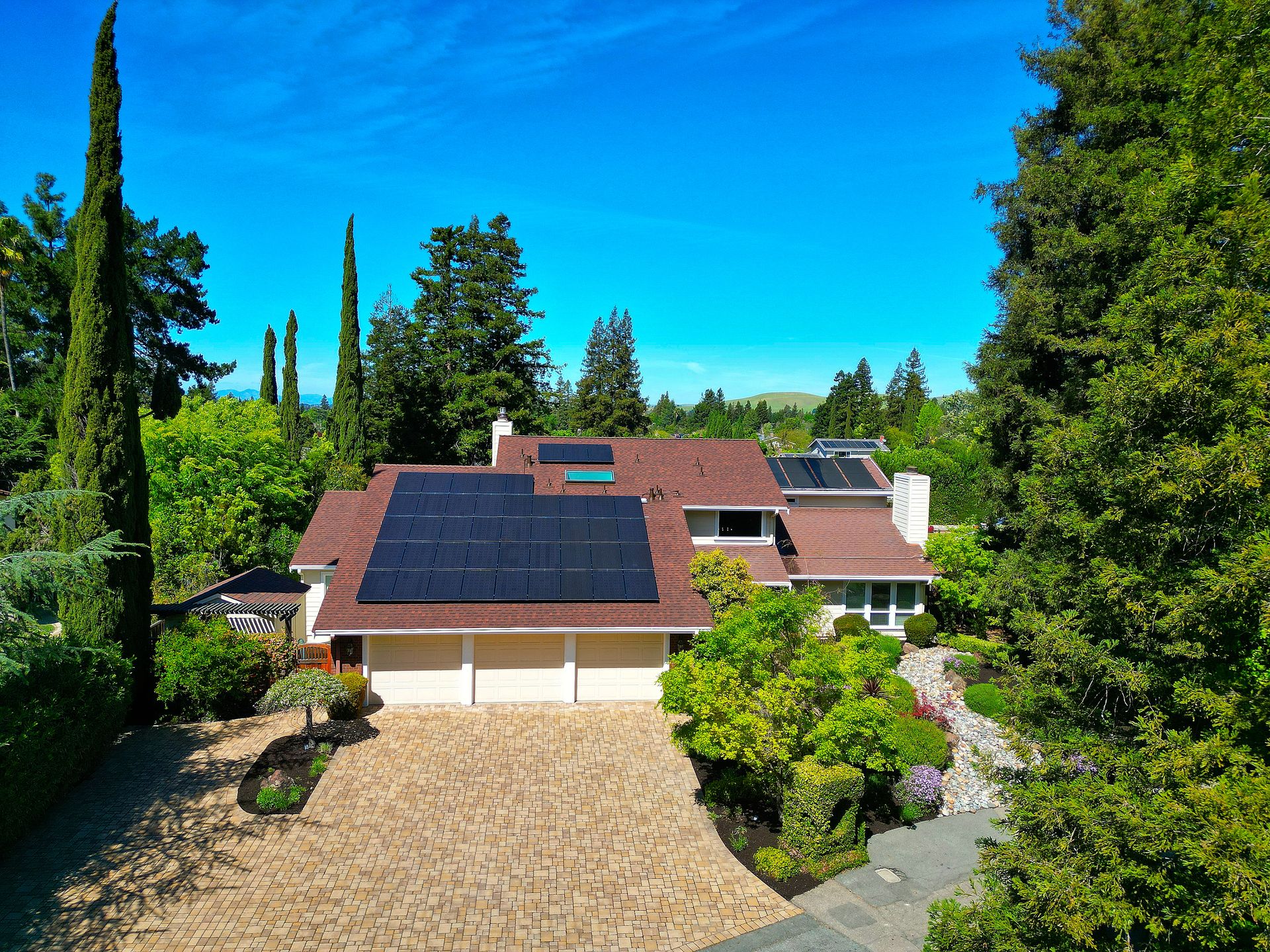 This elevated view captures a well-maintained, multi-level suburban home nestled among lush, mature greenery. The residence features a prominent brick-paved driveway leading to a three-car garage, complemented by extensive solar panel installations on the roof. The combination of manicured landscaping, stone elements, and a serene backyard setting conveys an inviting and established residential atmosphere.