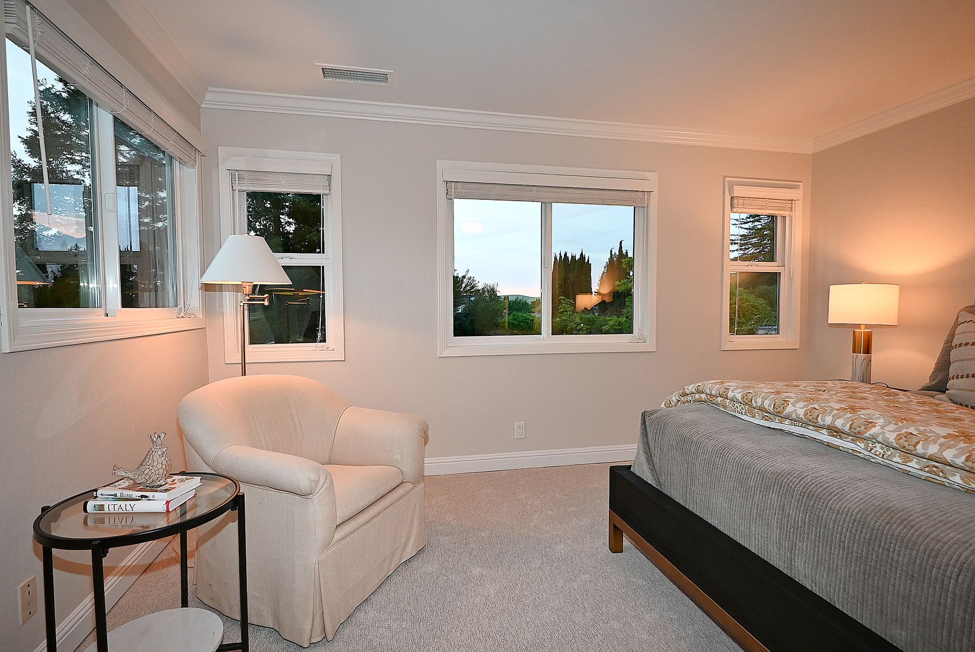 This tranquil primary bedroom features a comfortable beige armchair paired with a chic glass side table holding books and a decorative accent. The space is highlighted by neutral walls, plush carpeting, and a cozy bed with quality linens, all illuminated by warm table and floor lamps. Multiple large windows provide ample natural light and a pleasant view of the outdoor greenery.