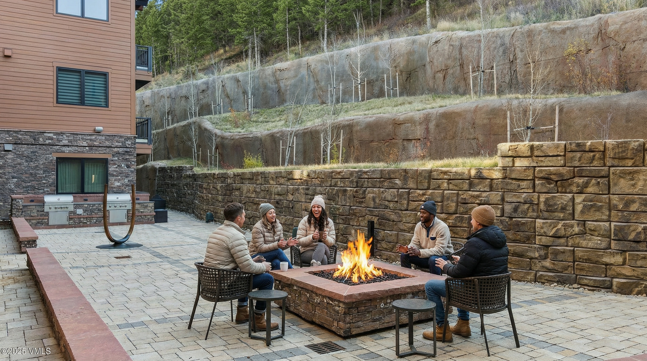This image showcases a communal patio area featuring a stone fire pit surrounded by chairs, set against a backdrop of a retaining wall and trees. The patio is paved with interlocking stones, presenting an inviting outdoor social space. It evokes a sense of community and relaxation, making it an attractive selling point.