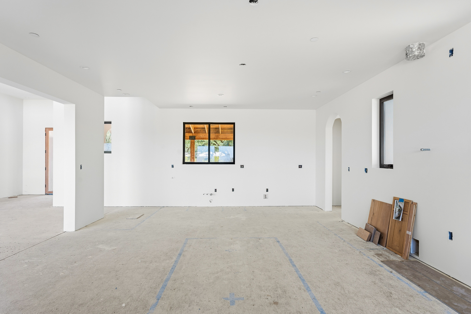 This interior view shows a kitchen under construction, currently featuring an open floor plan with freshly painted white walls and concrete flooring. The area exhibits clear layout markings for cabinets, a centralized window overlooking an outdoor structure, and an arched entryway that leads to another section of the home. This space presents a blank canvas with potential for a modern custom design.
