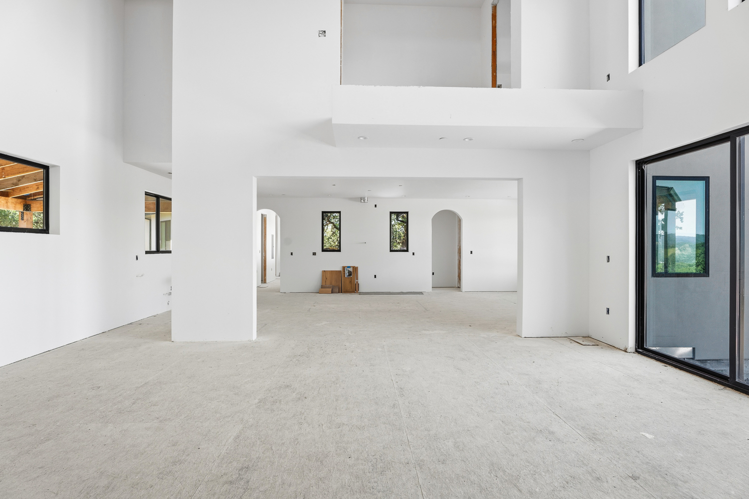 This spacious, modern open-concept living area is currently in the construction phase, showcasing high ceilings, crisp white walls, and smooth concrete flooring. The design features large black-framed windows and arched transitions that provide architectural interest and connection between spaces. This room offers a bright, minimalist canvas ready for interior finishing touches.