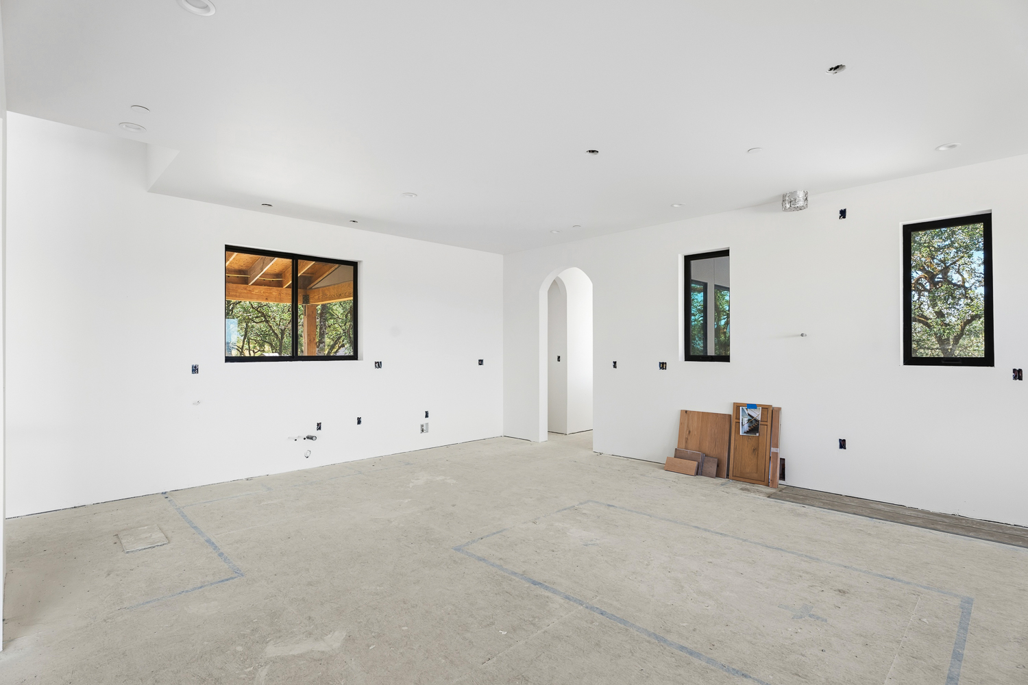 This image depicts an unfinished interior space, likely intended for a kitchen remodel, characterized by its open-concept layout, white walls, and pre-wired electrical points for appliances and lighting. The room features a prominent arched doorway, large modern windows with dark frames, and floor markings suggesting the future placement of cabinetry or an island. The space offers a blank canvas with a neutral palette, highlighting its potential for a custom design.