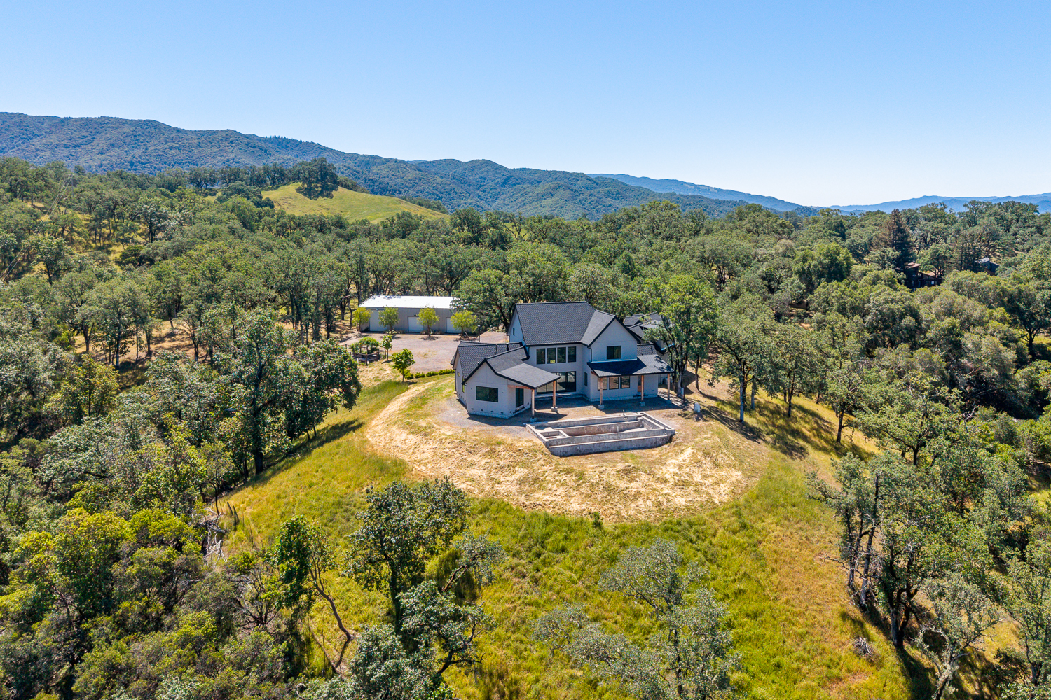This stunning aerial view captures a contemporary farmhouse-style residence situated prominently atop a rolling, tree-studded hilltop. The property features a multi-gabled home with a large, unfinished swimming pool structure, surrounded by expansive grassy terrain and mature oak forests. The setting offers a serene, private atmosphere with far-reaching mountain views in a picturesque rural landscape.