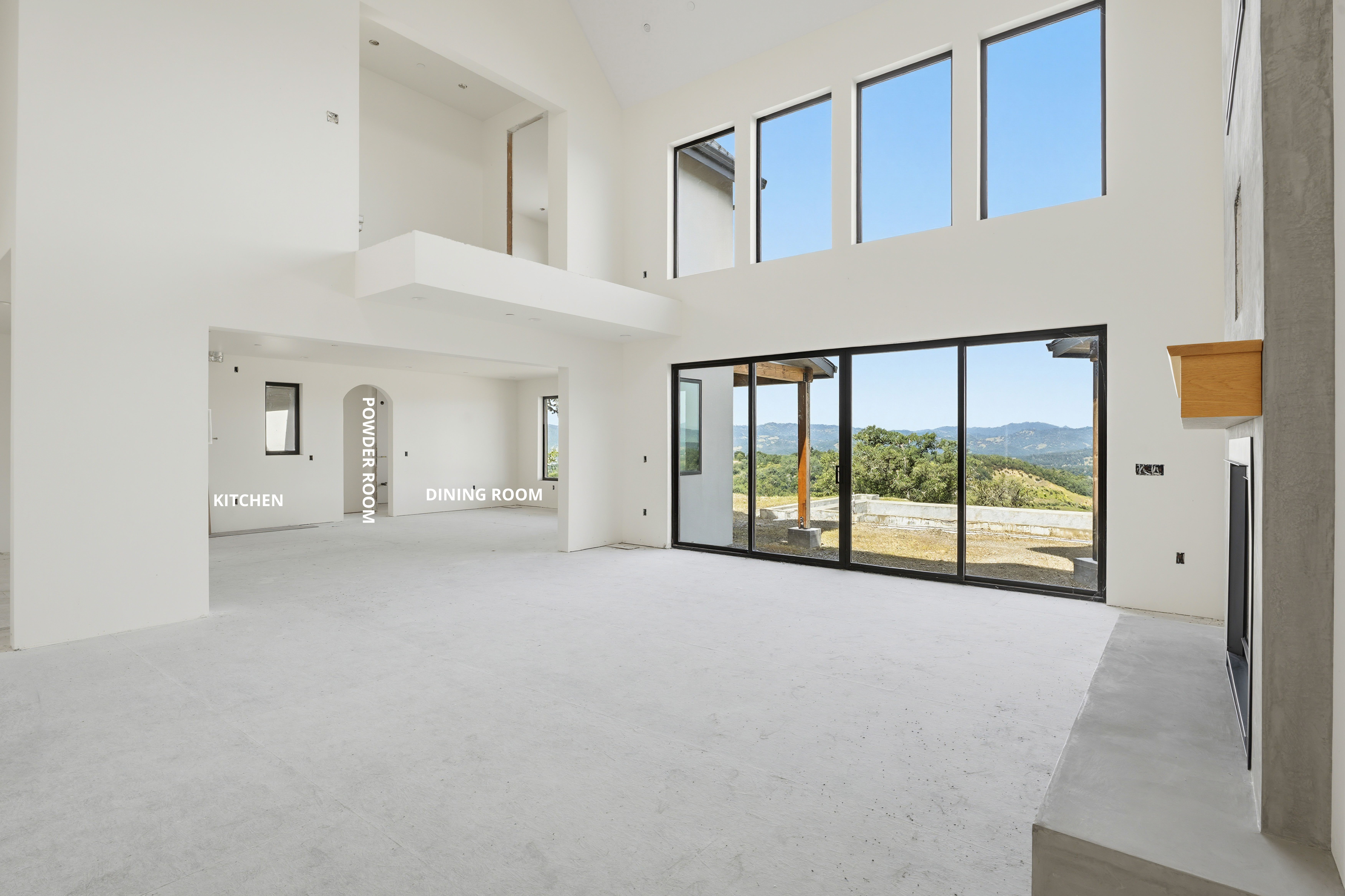 This spacious, double-height living room features an open, minimalist design with smooth white walls and a large, concrete-hearth fireplace with a wooden mantel. A massive floor-to-ceiling glass wall provides an expansive view of the surrounding hills, while adjacent transitions lead towards the kitchen, dining room, and a powder room. The neutral foundation and natural light create a modern, airy atmosphere ready for customization.