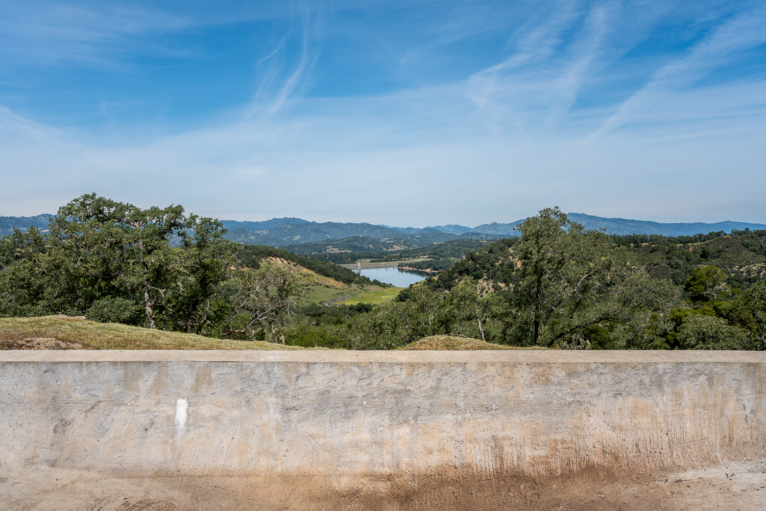This image showcases a scenic, expansive view overlooking rolling hills, scattered oak trees, and a distant reservoir. A concrete wall in the foreground frames the landscape, emphasizing the natural beauty and elevated vantage point of the property. The setting offers a serene, panoramic outlook, ideal for showcasing the picturesque surroundings of a rural or semi-rural estate.