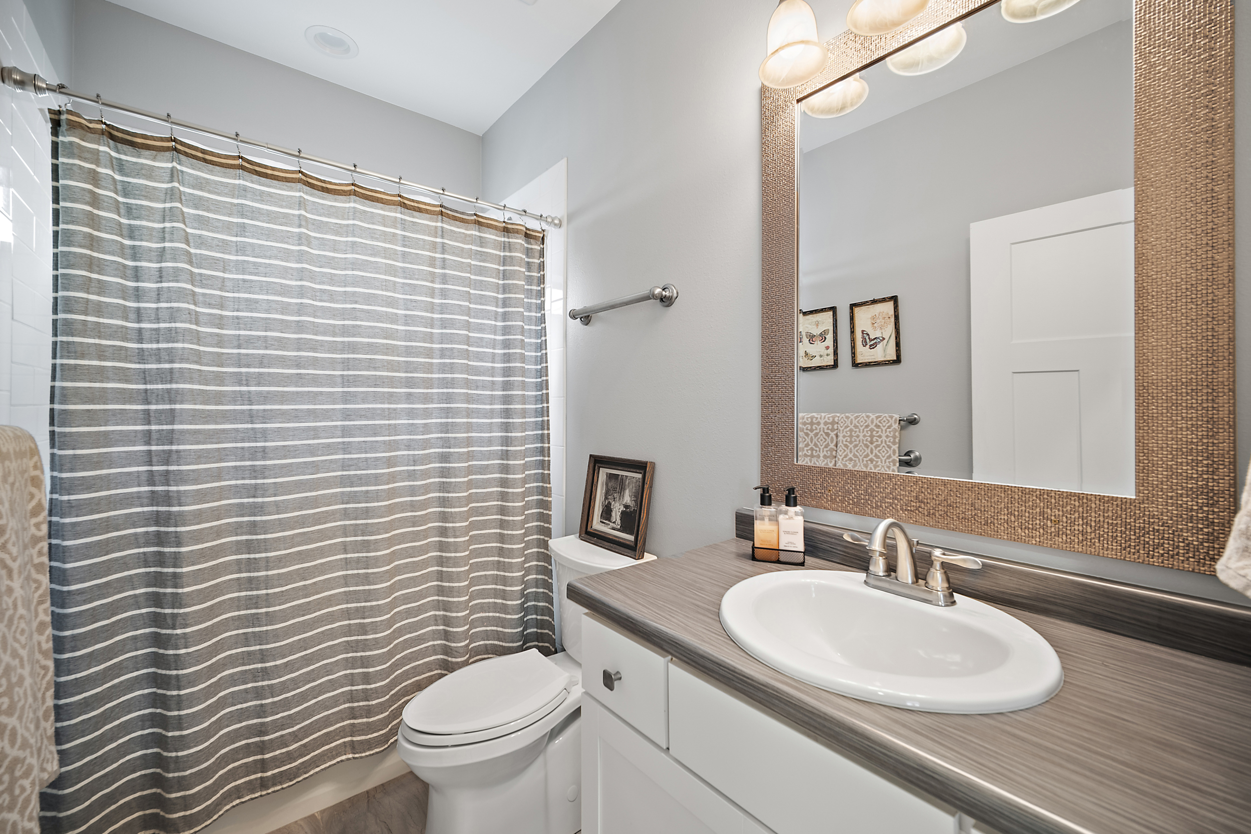 This clean and functional bathroom features a neutral color palette with light gray walls and white cabinetry. The space includes a framed vanity mirror, a modern sink, and a shower-tub combination with a striped curtain for a calm, contemporary aesthetic. Small decorative touches like framed art and a picture on top of the toilet add a personalized, homey feel.
